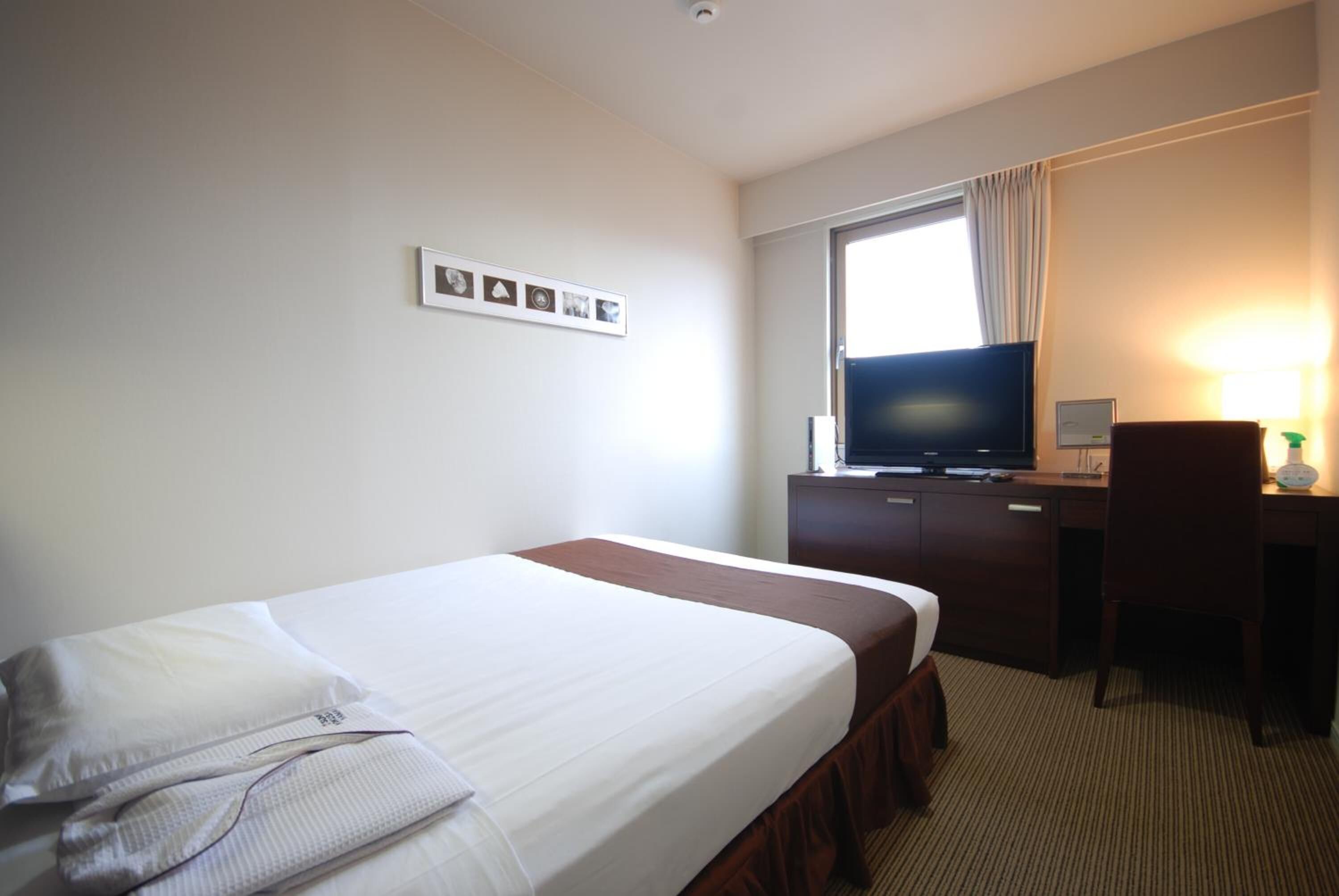 Quarto de hotel com cama, escrivaninha, televisão e janela com cortinas.