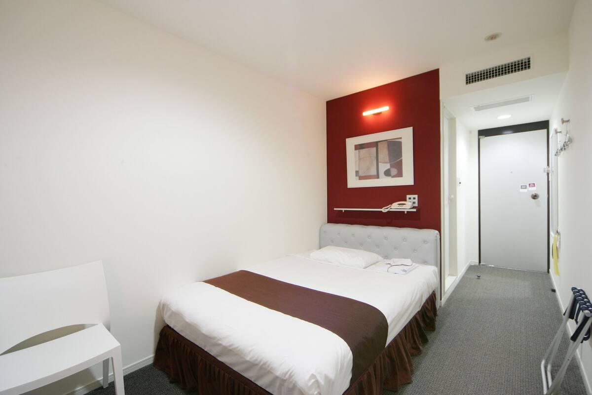 Une chambre d’hôtel avec un lit, une chaise blanche, un mur d’accent rouge et une porte.