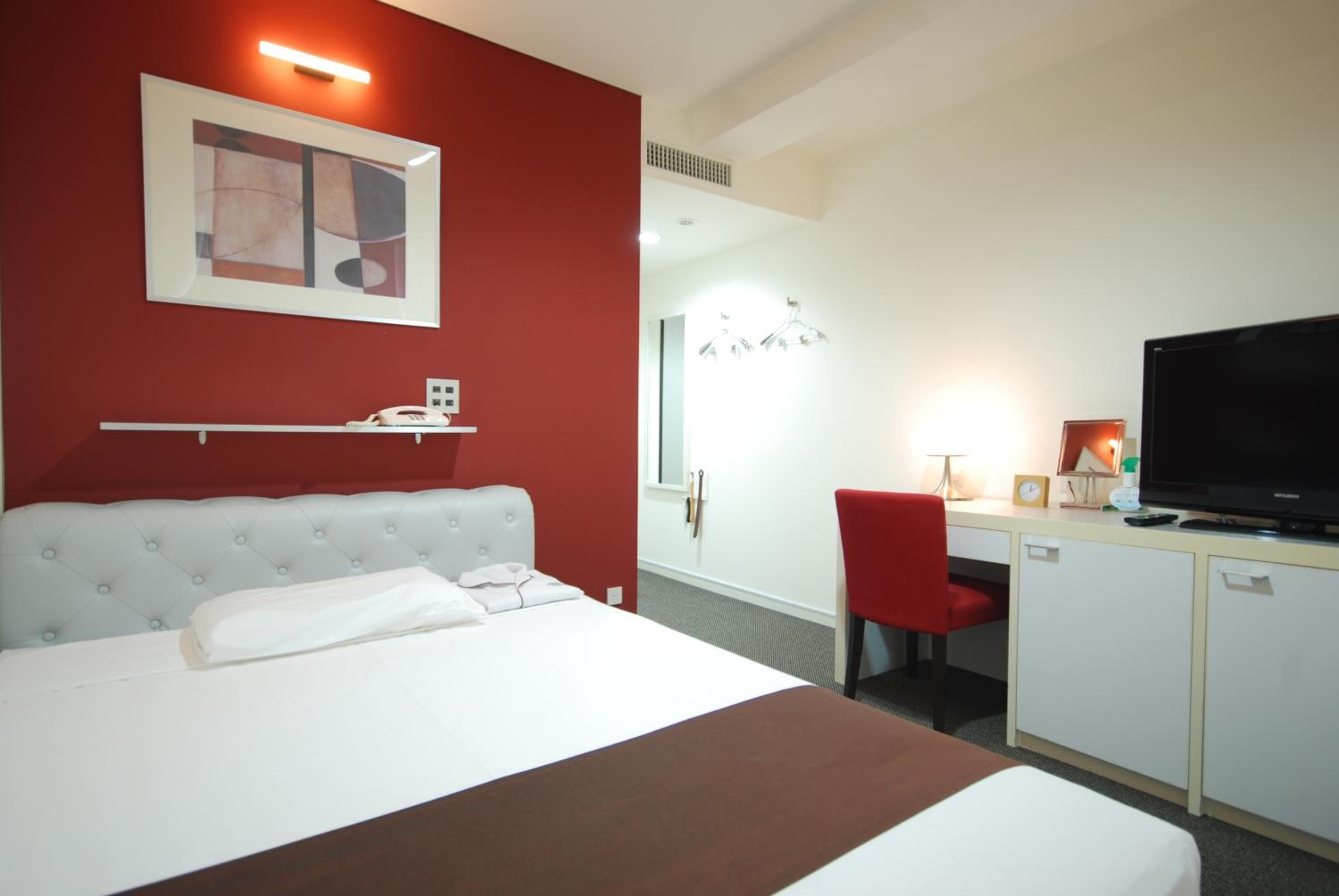 A hotel room with a bed, a desk with a TV, a red chair, and a red wall.