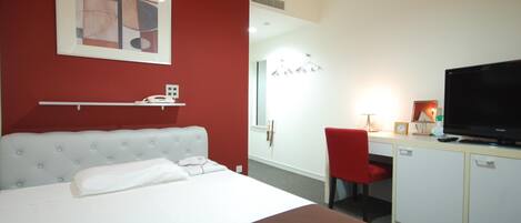 A hotel room with a bed, a desk with a TV, a red chair, and a red wall.