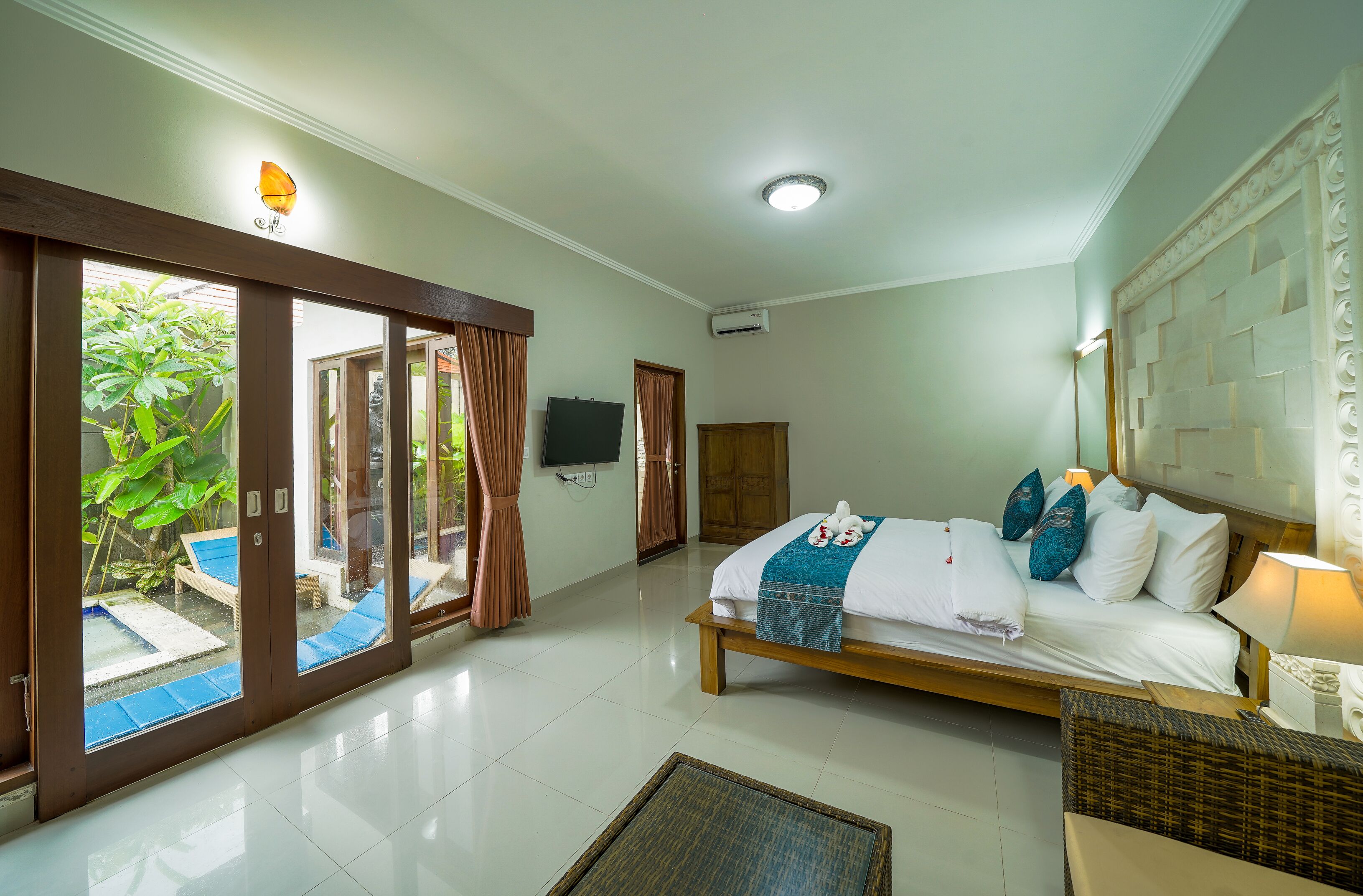 A hotel room with a bed, a TV, a window with curtains, and a view of greenery outside.