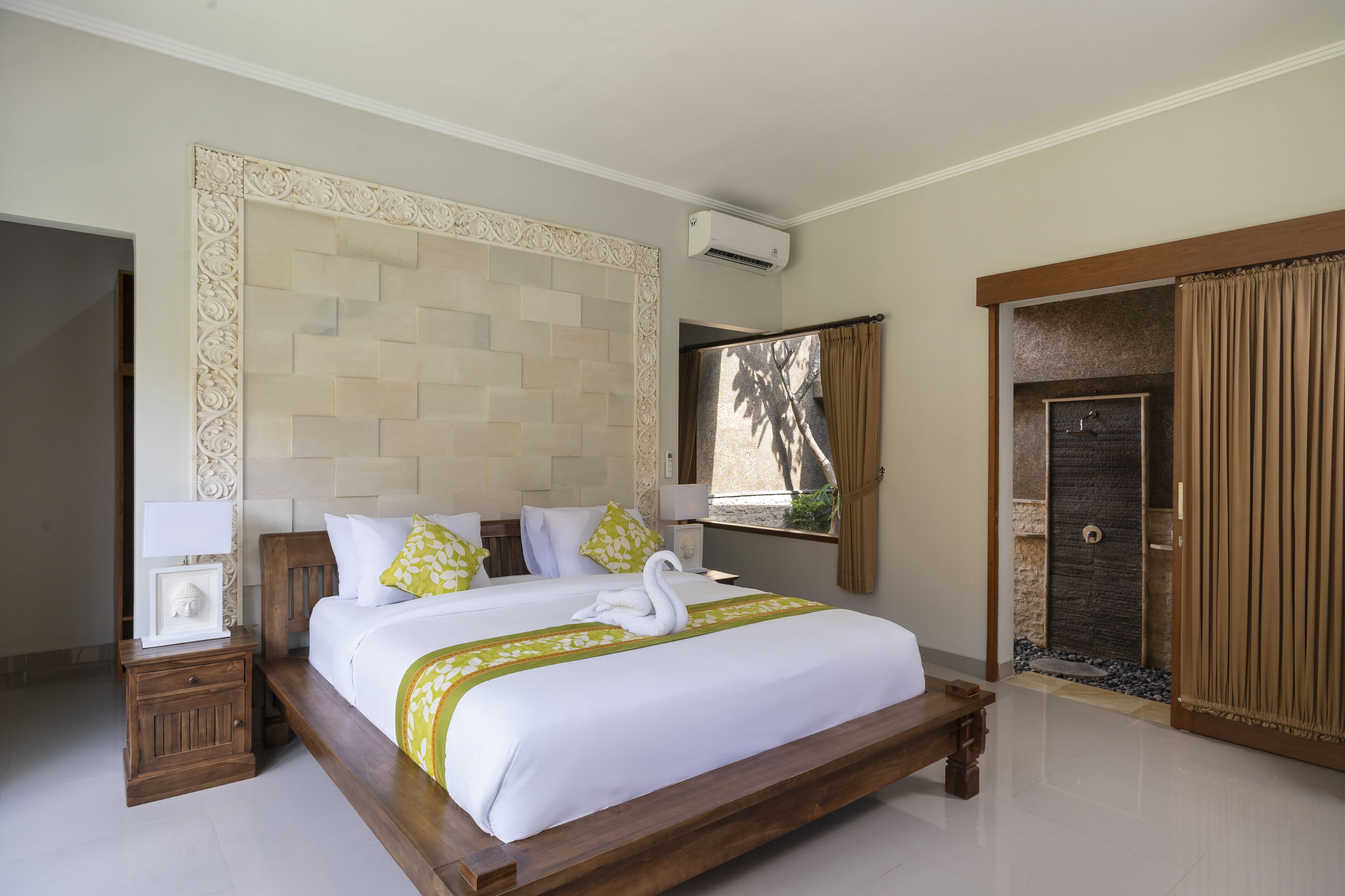 A hotel room with a large bed, a wooden headboard, a nightstand, a window with curtains, and a door with a view of the outside.