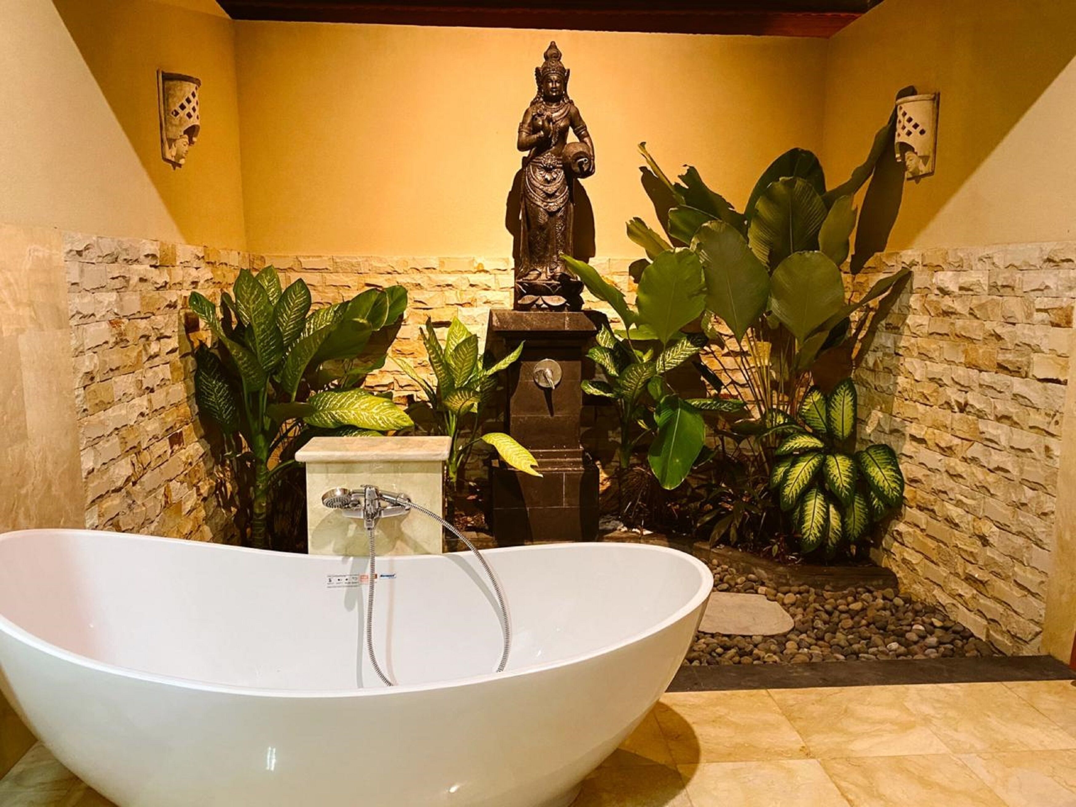 A bathroom with a freestanding bathtub, a statue, and tropical plants.