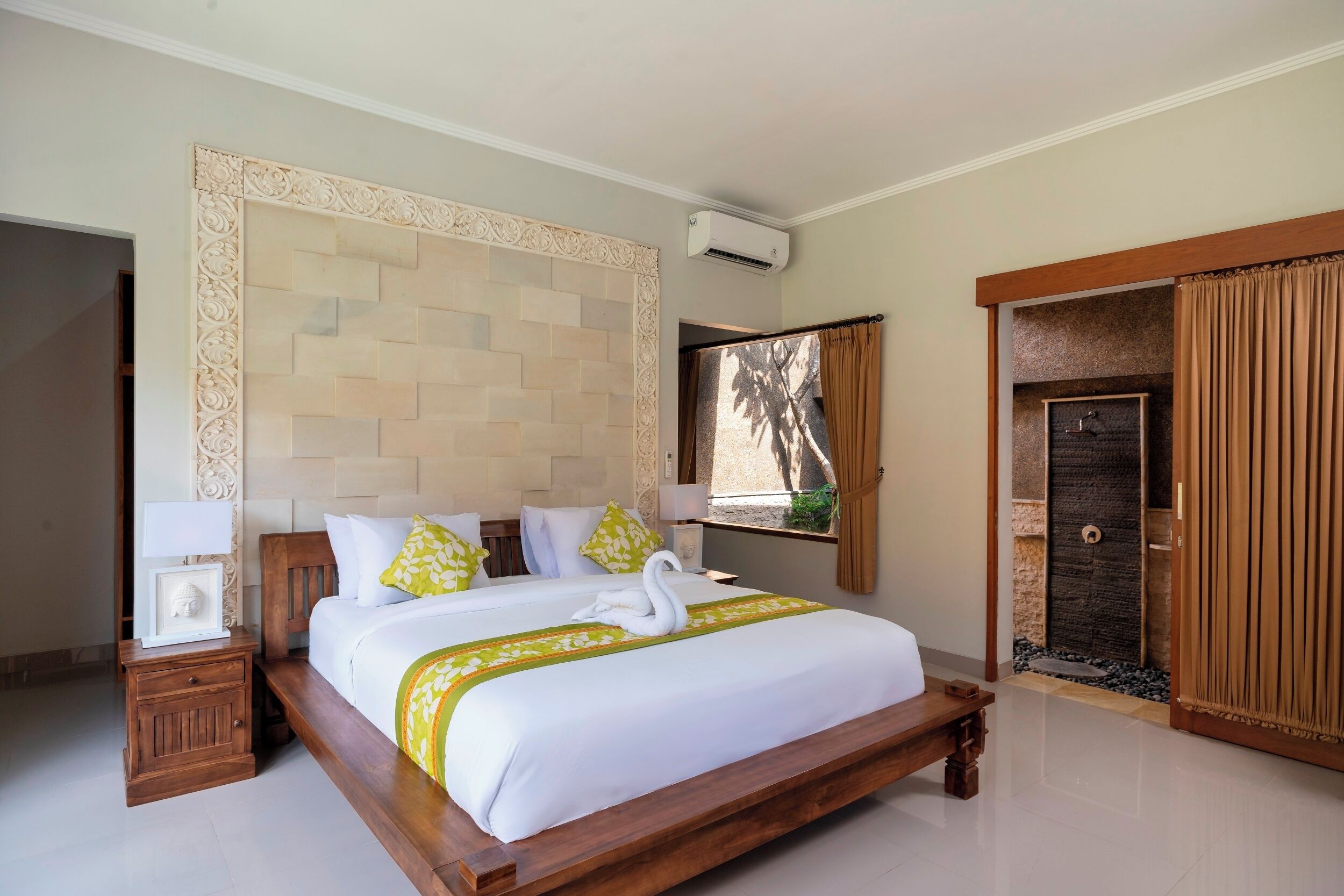 Une chambre d’hôtel avec un grand lit, une tête de lit en bois, une table de chevet avec une lampe, une fenêtre avec des rideaux et une porte avec un cadre en bois.