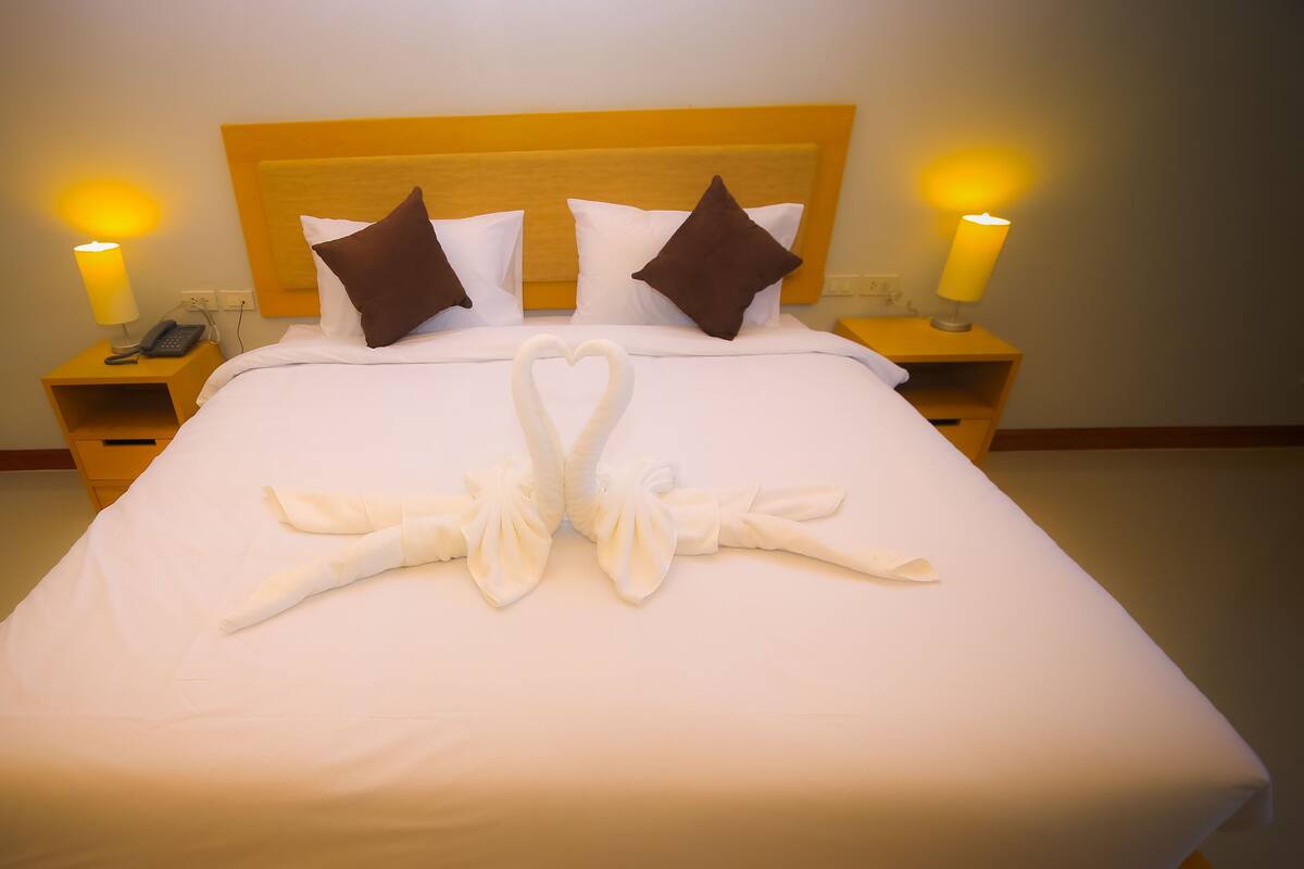 A neatly made bed with a heart-shaped towel arrangement, a wooden headboard, and bedside lamps.