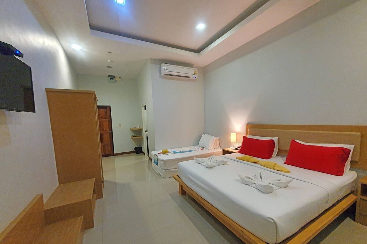 A hotel room with a large bed, bedside tables, a TV, an air conditioning unit, and a wooden bench.