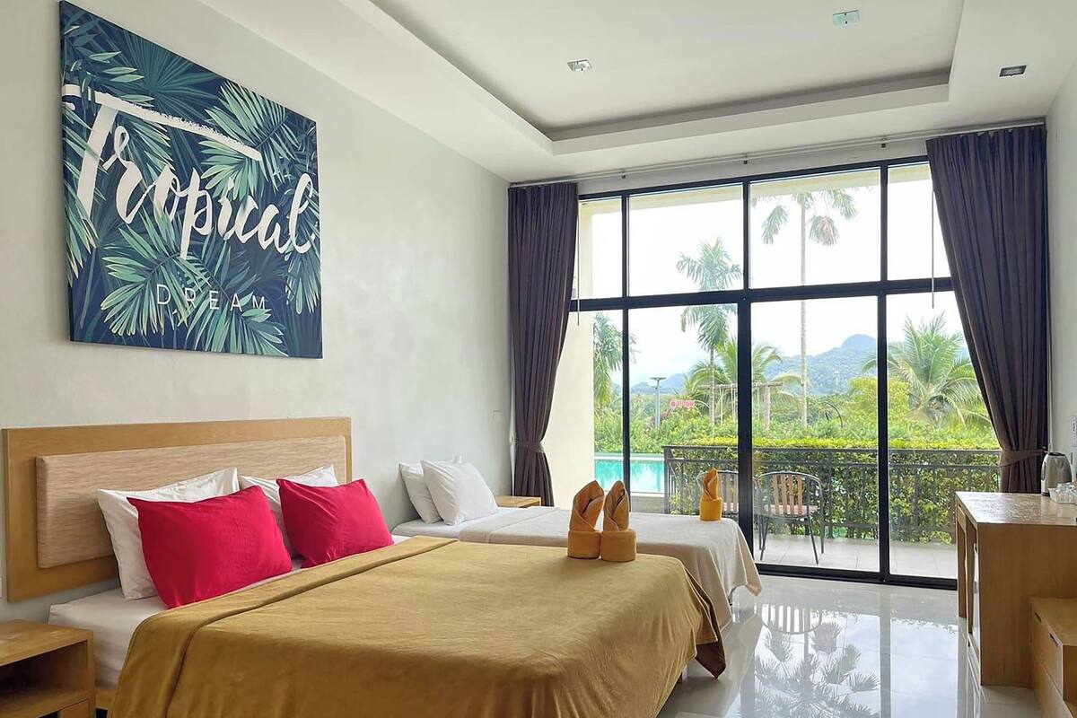 A hotel room with a large window, a bed with a yellow bedspread, a wooden headboard, and a tropical-themed wall art.
