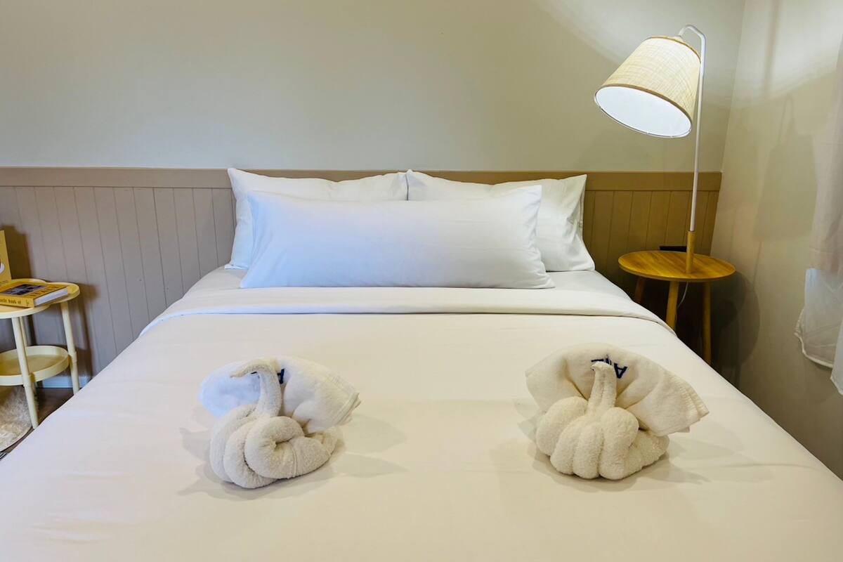 A hotel room with a neatly made bed, two towels shaped like swans, a bedside lamp, a small wooden table, and a chair.