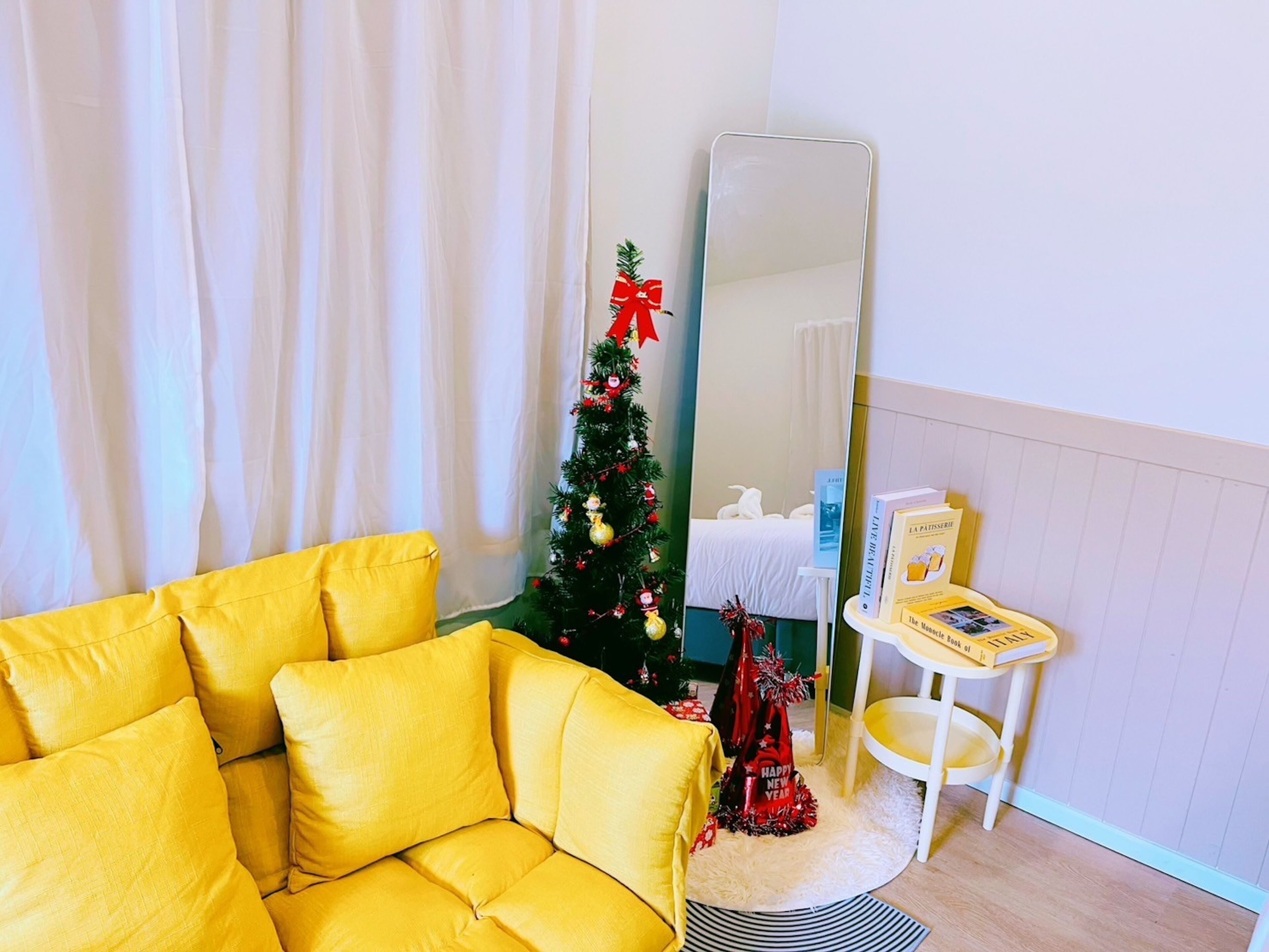A room with a yellow sofa, a Christmas tree, a mirror, and a small table with books.
