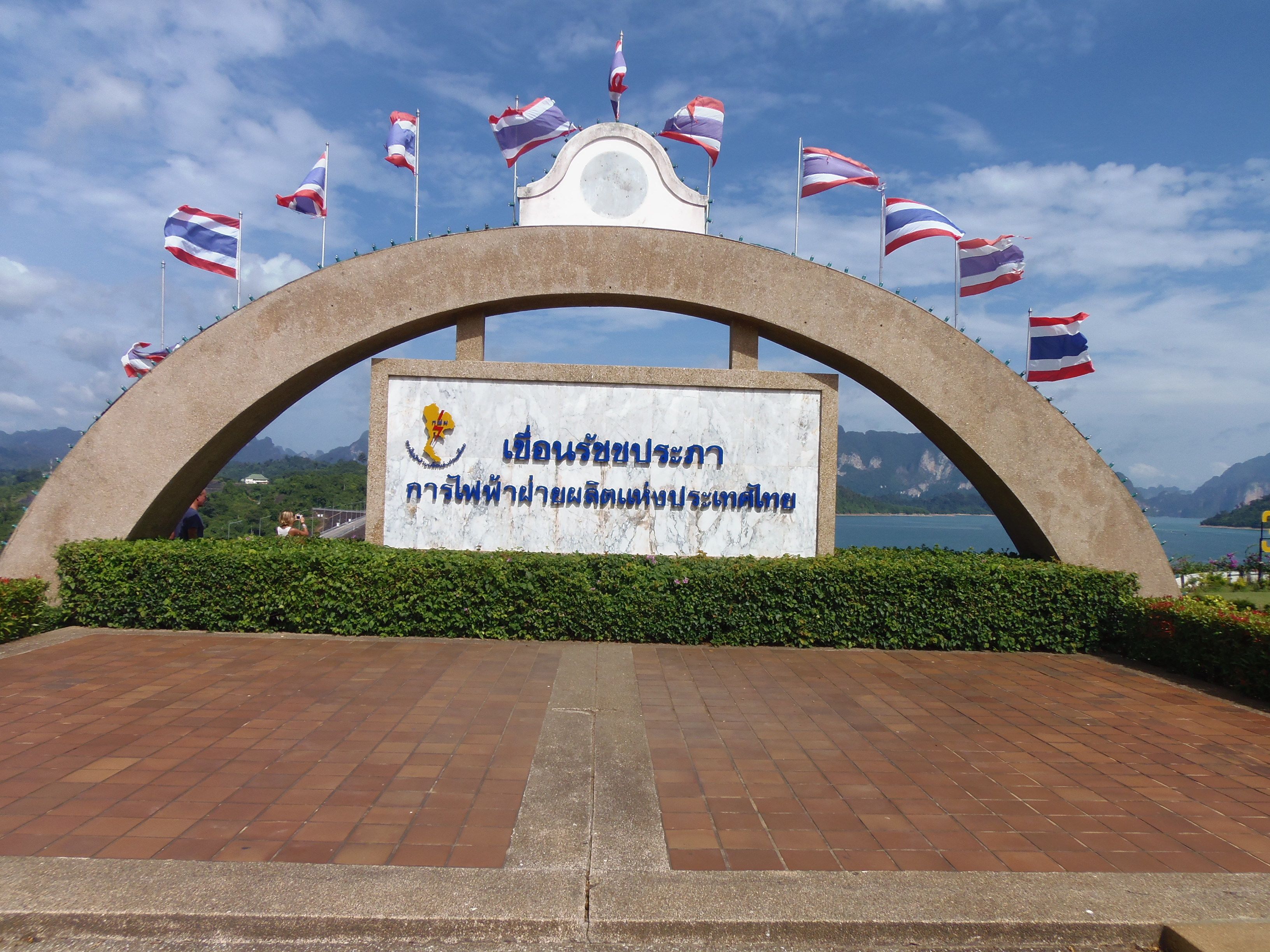 A large archway with a sign in front, surrounded by flags and a scenic view of mountains and water.