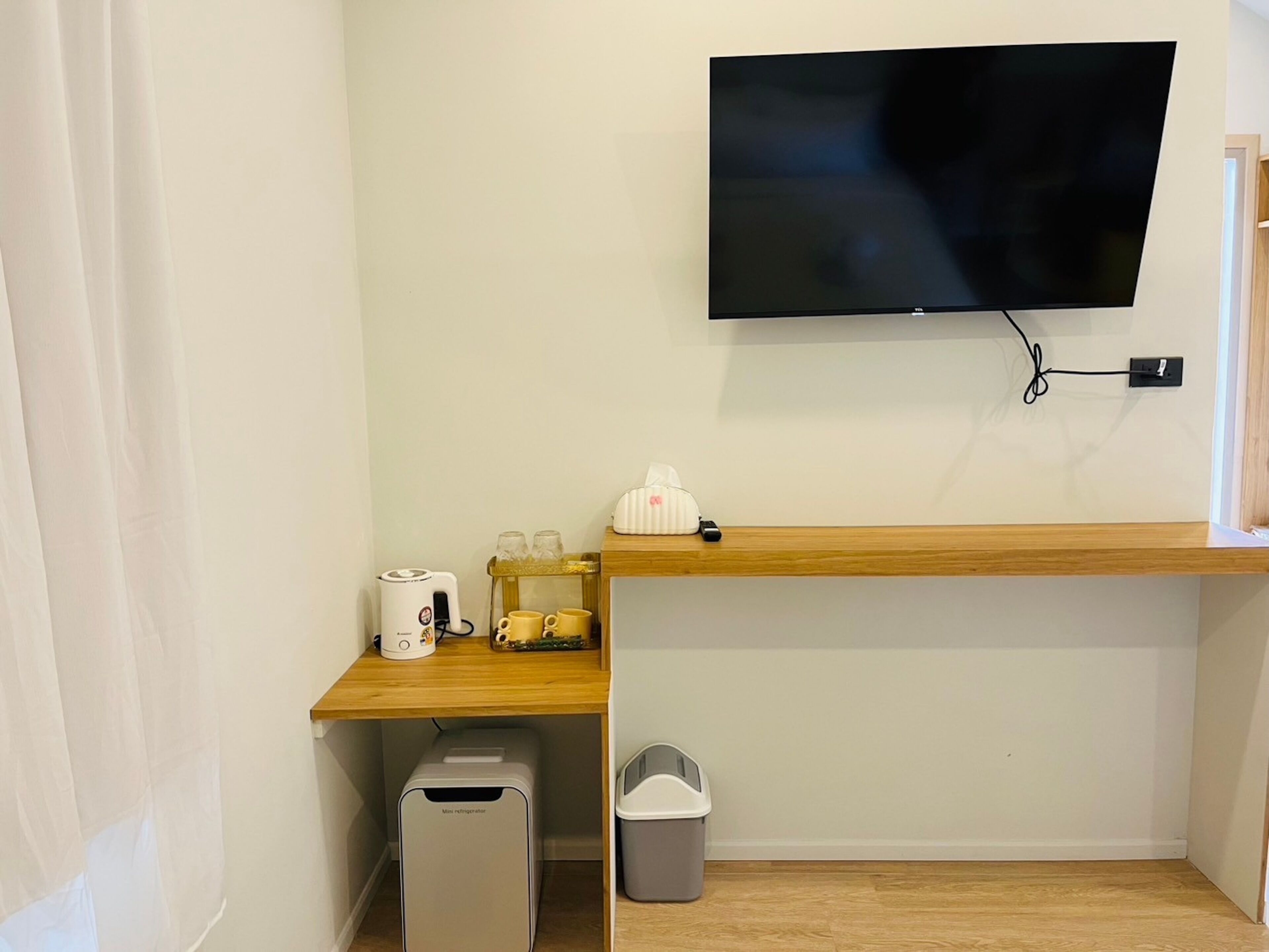 Una stanza piccola con una TV montata a parete, una scrivania in legno con un bollitore, tazze e un box di fazzoletti. C'è anche un mini frigorifero e un cestino.
