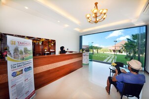 Zona de recepción de un hotel con un mostrador de madera, un candelabro y un amplio ventanal que ofrece vistas a un césped cuidado y edificios al fondo.