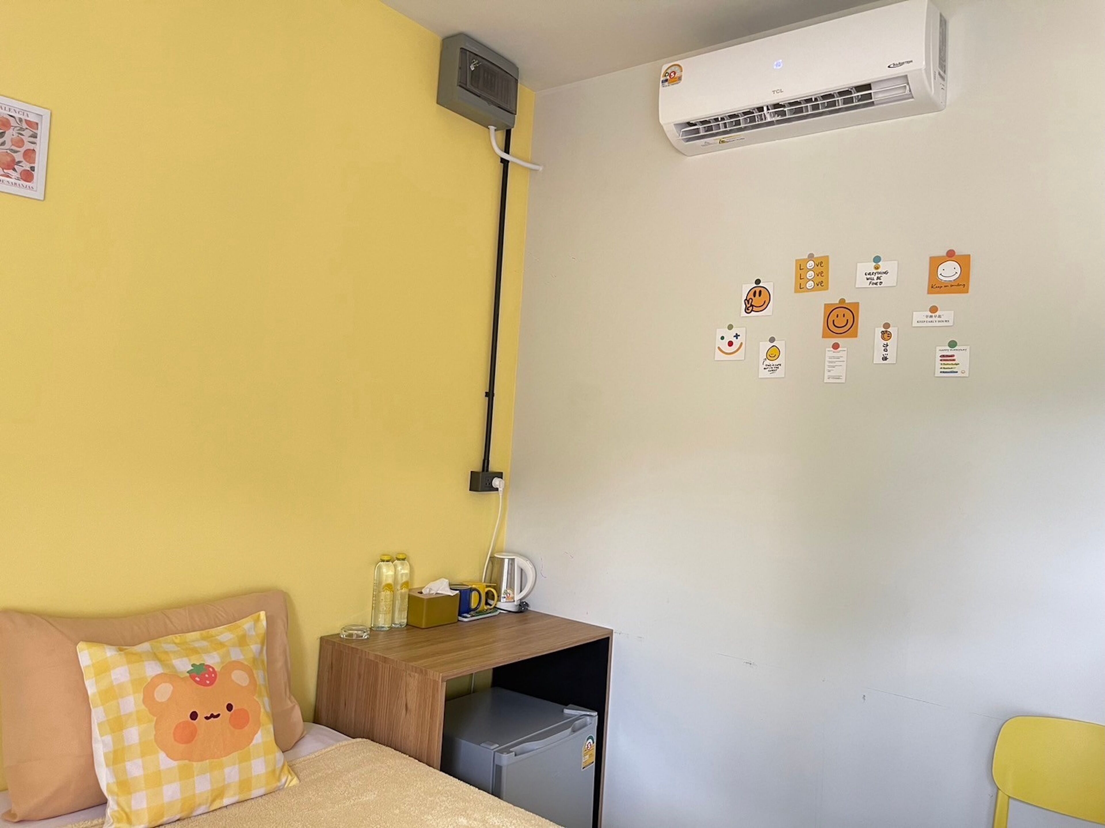 A room with a yellow wall, a bed with a checkered pillow, a small wooden table with a kettle, and a wall-mounted air conditioner.