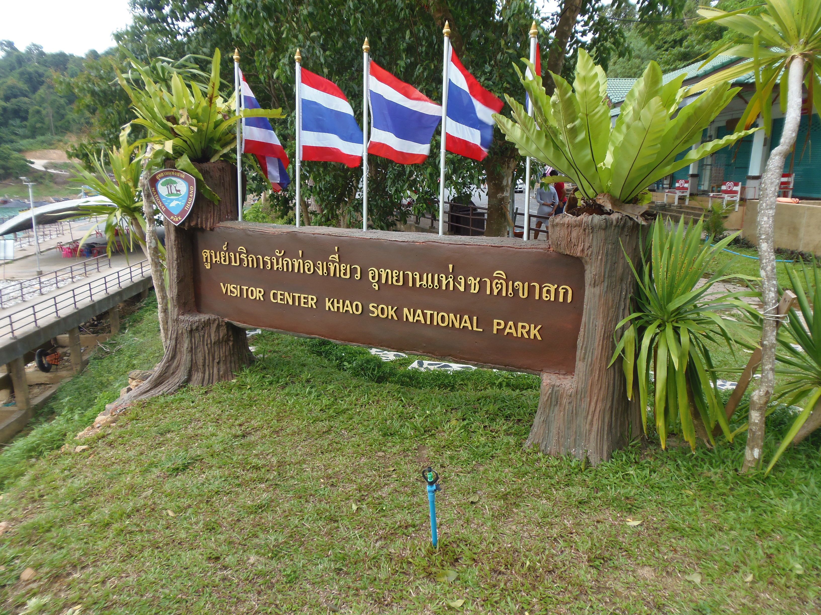 Naamplaatje voor het bezoekerscentrum in het Khao Sok Nationaal Park, met Thaise en Engelse tekst, omringd door vlaggen en tropische planten.