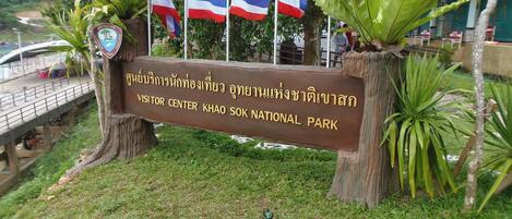Naamplaatje voor het bezoekerscentrum in het Khao Sok Nationaal Park, met Thaise en Engelse tekst, omringd door vlaggen en tropische planten.