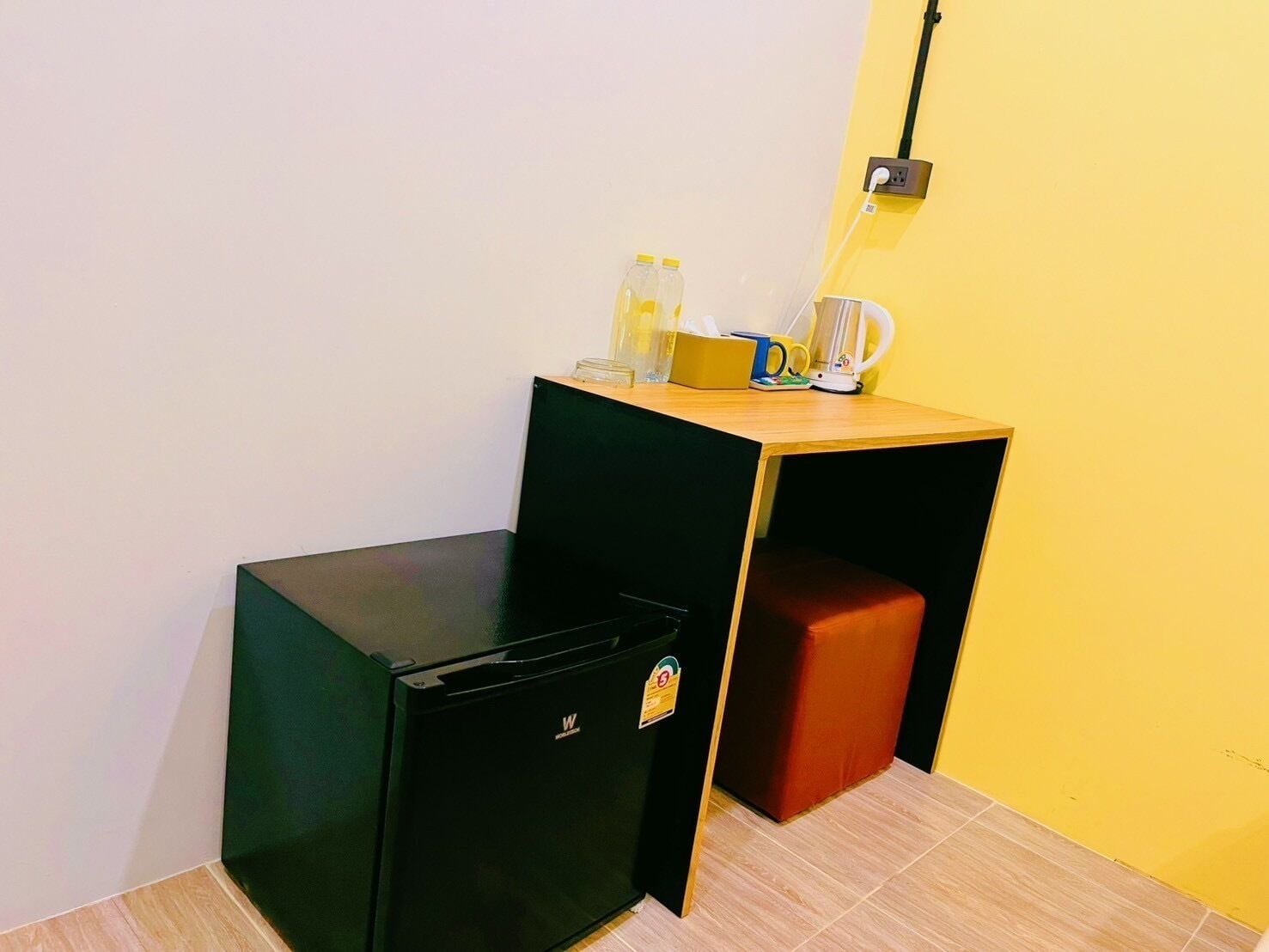 Une pièce de taille modeste, avec un petit réfrigérateur noir, un bureau en bois devant un mur jaune et un banc rouge.