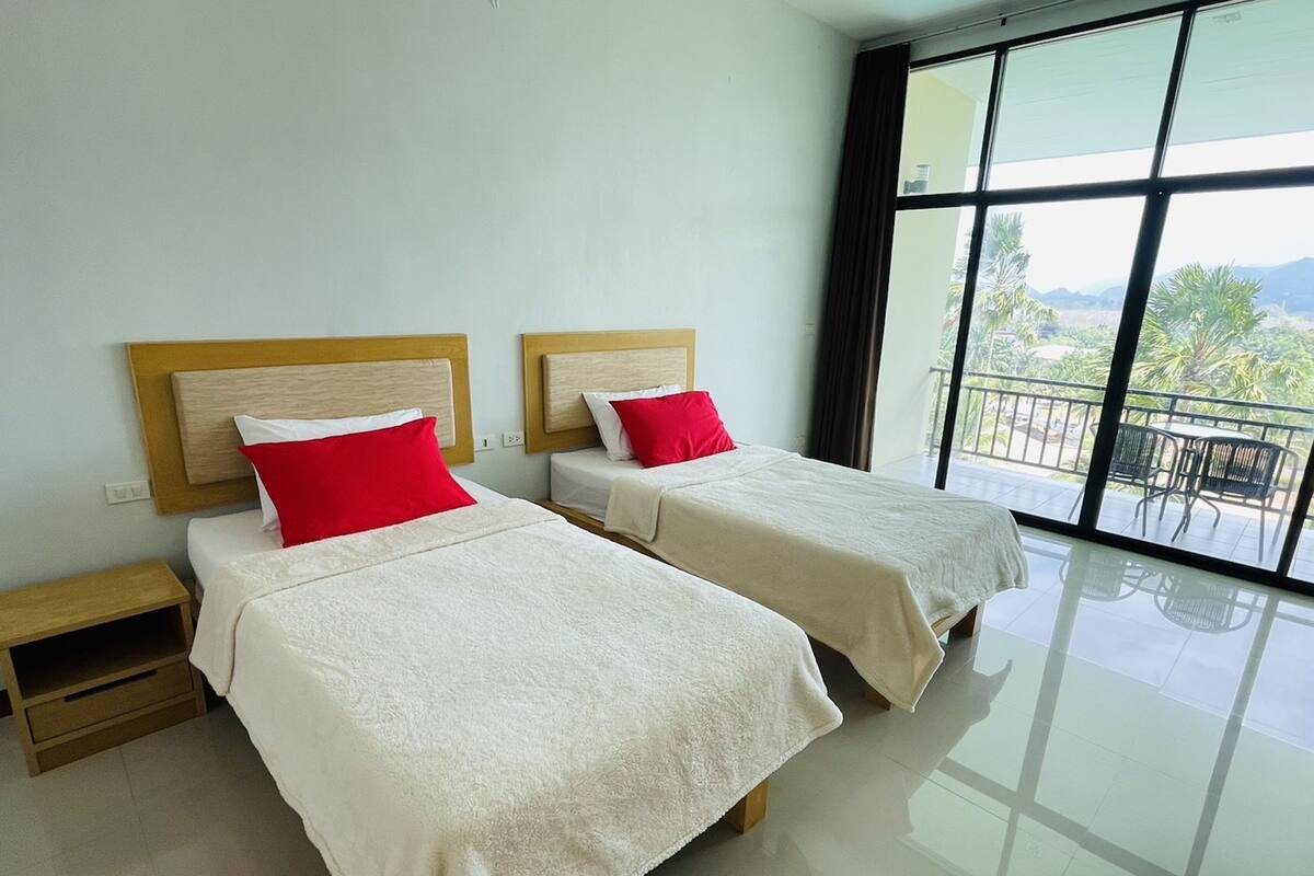A hotel room with two beds, each with a wooden headboard and red pillows. The room has a large window offering a view of palm trees and mountains.