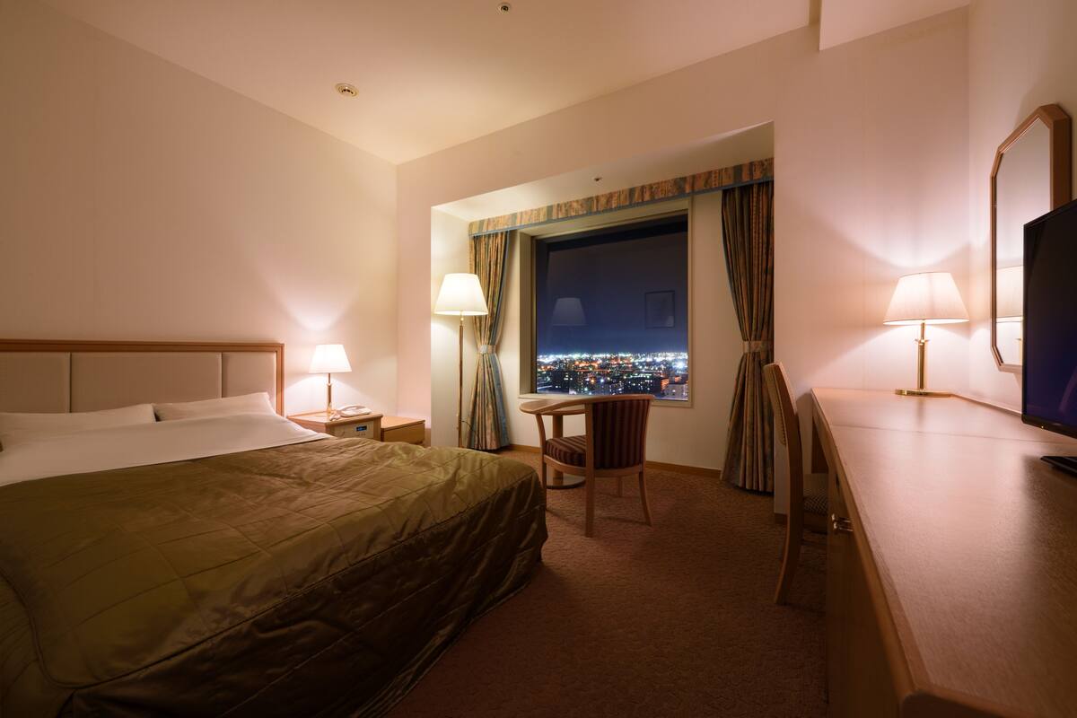 A hotel room with a bed, a desk, a chair, a TV, and a window with a city view.