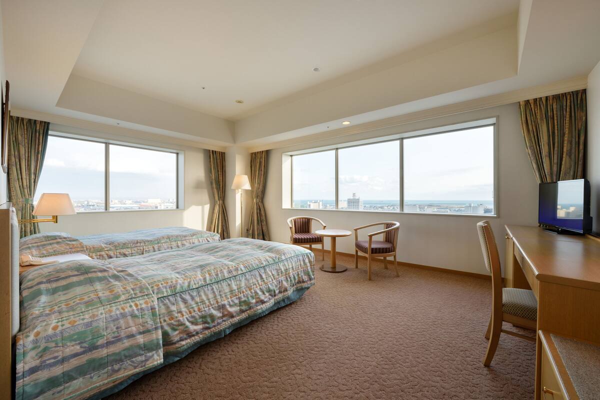 A hotel room with two beds, a desk, a chair, a TV, and large windows offering a city view.