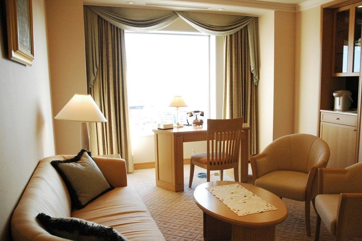 A hotel room with a sofa, a dining table, a chair, a lamp, and a view of the city.