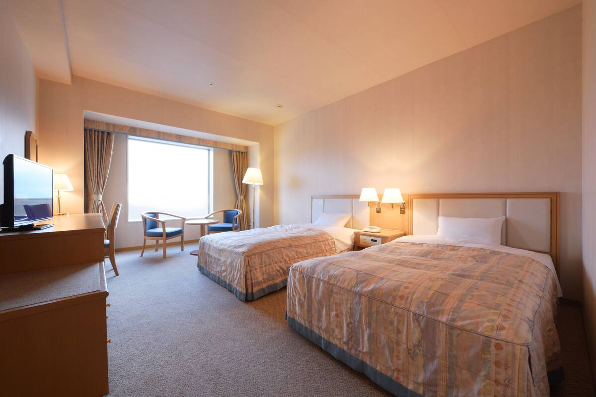 A hotel room with two beds, a desk with a computer, a chair, and a window with curtains.