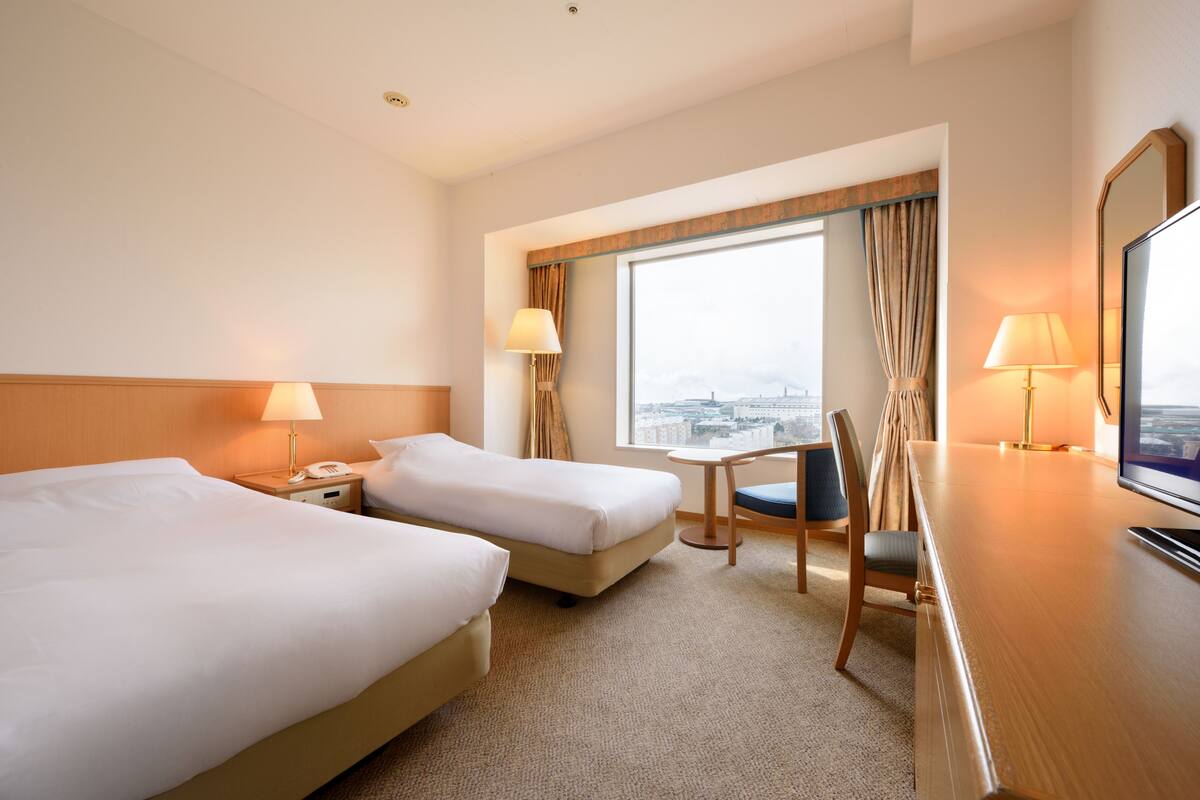 A hotel room with two beds, a desk, a chair, and a large window offering a city view.