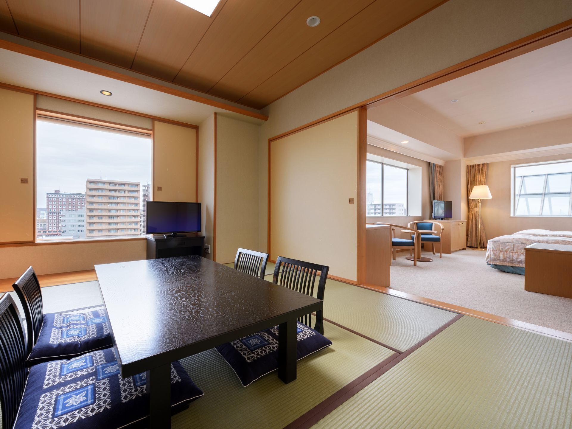 Quarto de hotel com mesa de jantar, cadeiras, televisão, cama e vista para a paisagem urbana.