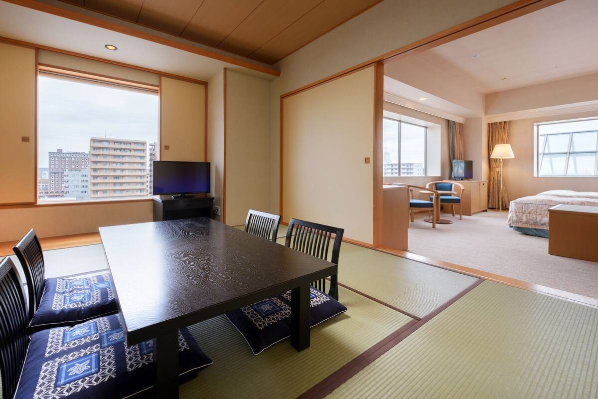A hotel room with a dining table, chairs, a TV, a bed, and a view of the cityscape.