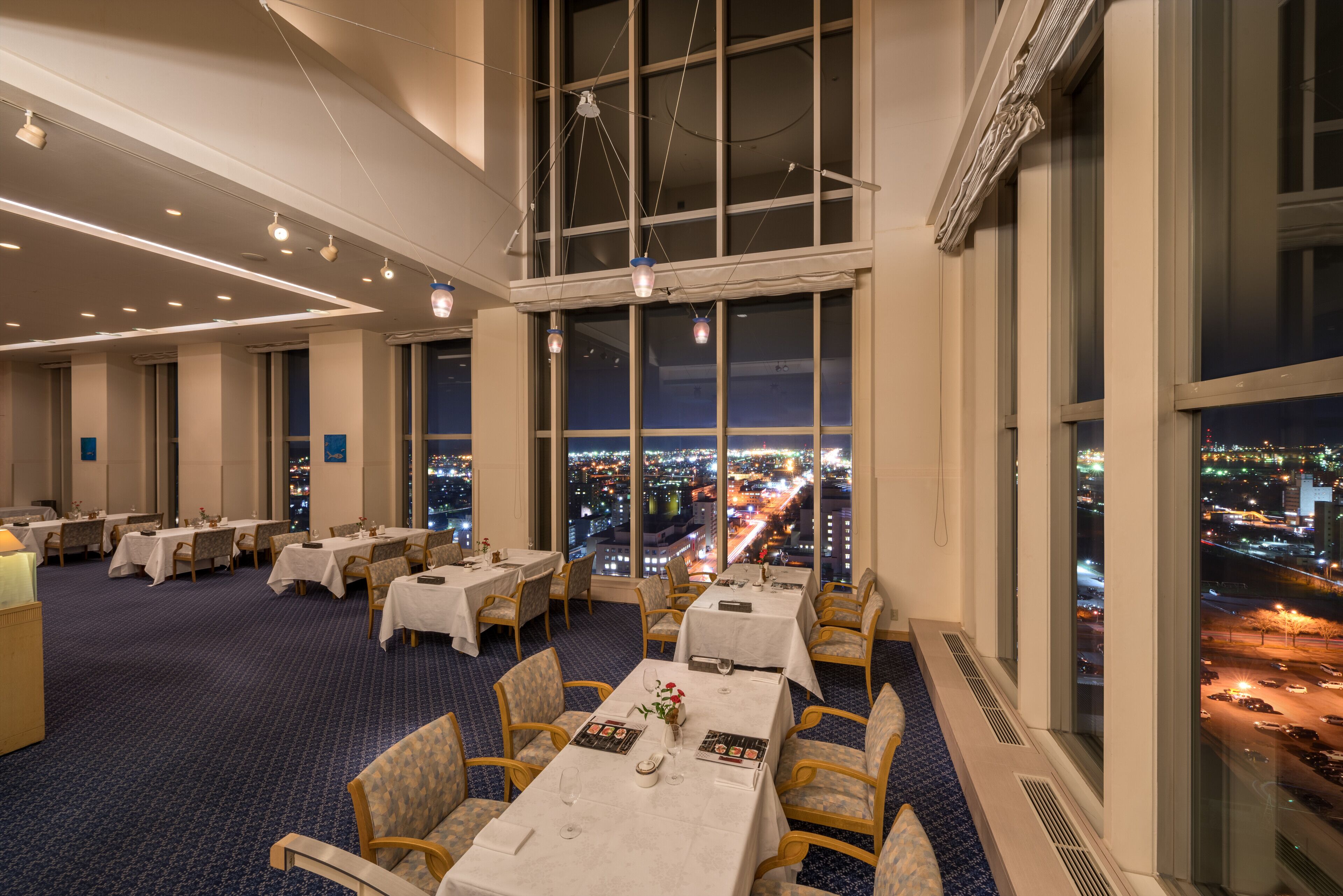 Uma área de refeições de hotel com mesas preparadas para hóspedes, grandes janelas com vista para a cidade e teto alto com luminárias modernas.