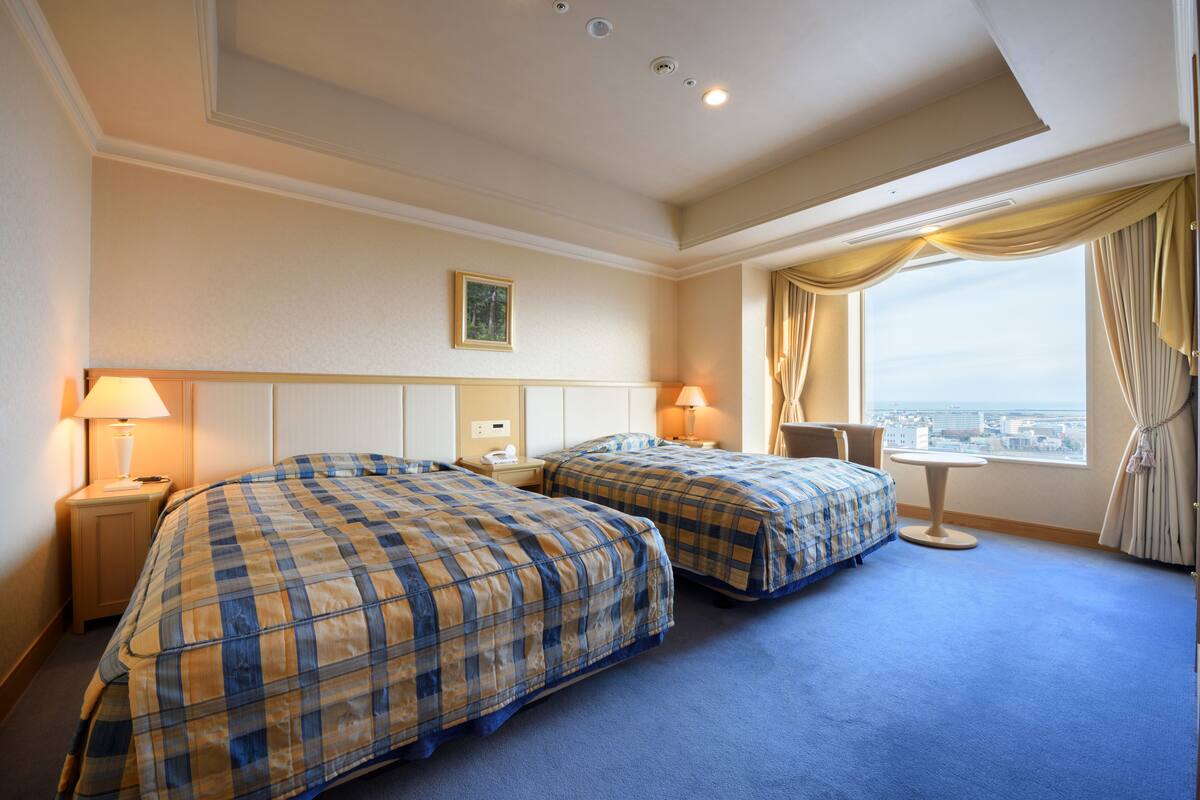 A hotel room with two beds, a small table, a chair, and a window with curtains.
