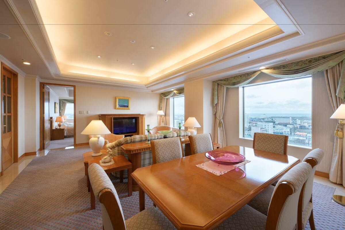 A hotel room with a dining area, a sofa, a television, and a view of the city.