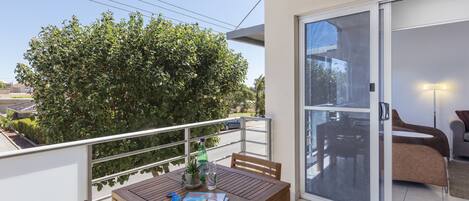 A balcony with a wooden table, chairs, and a magazine. There is a sliding glass door leading to an indoor living area with a sofa and a lamp.
