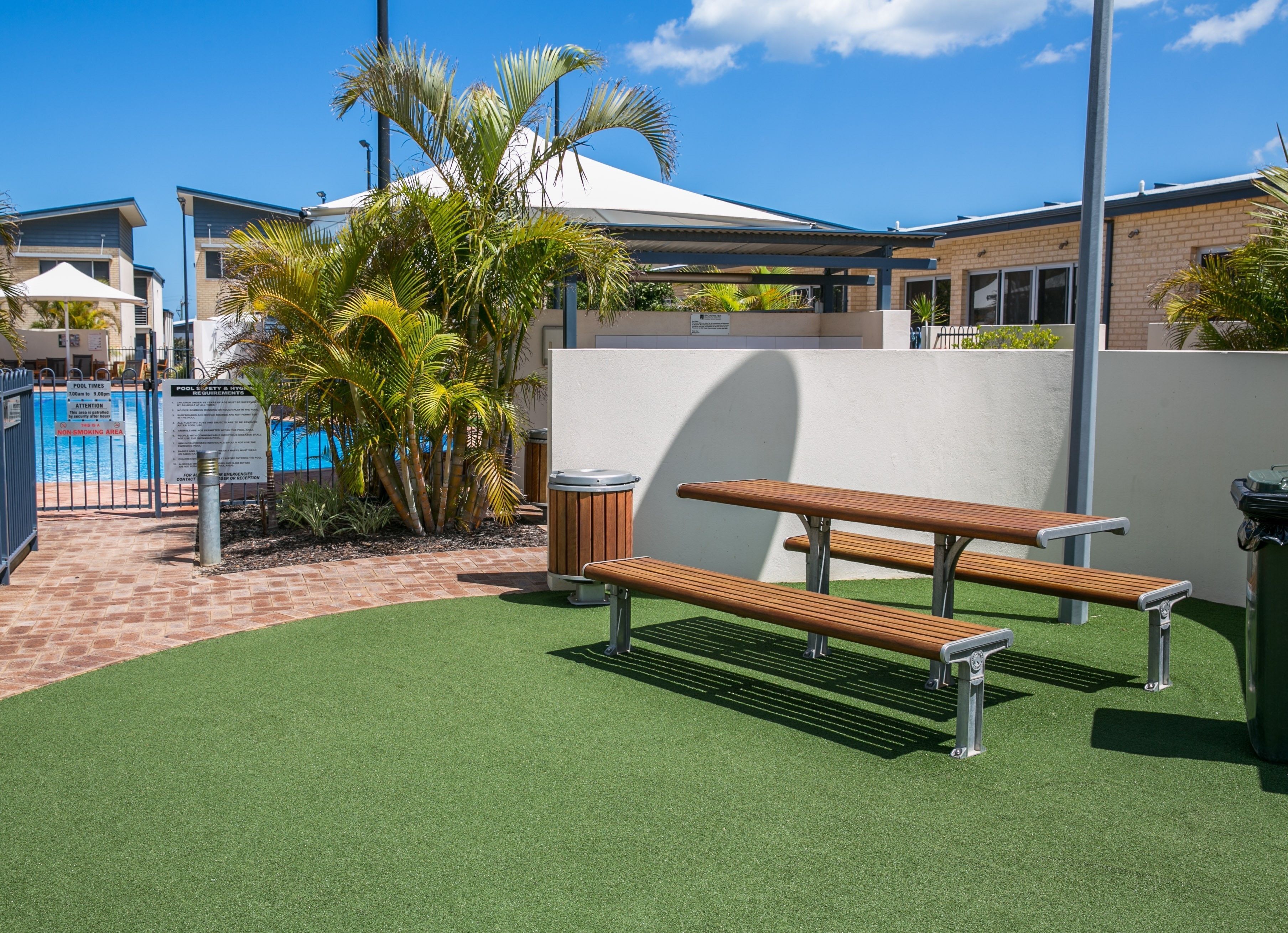 A poolside area with artificial turf, a wooden bench, a trash bin, and a white wall.