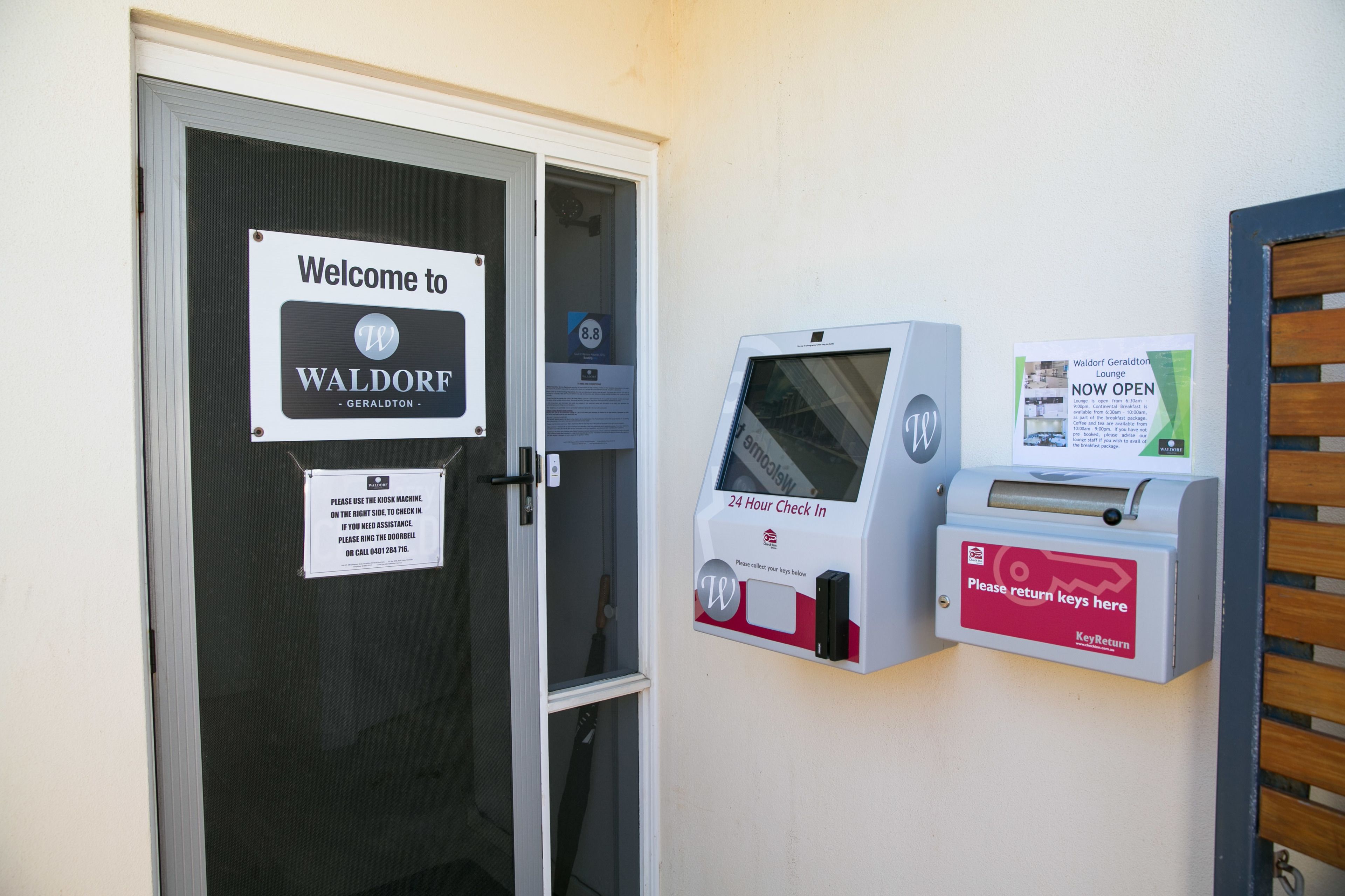 A hotel entrance with a welcome sign, a 24-hour check-in machine, and a key return box.