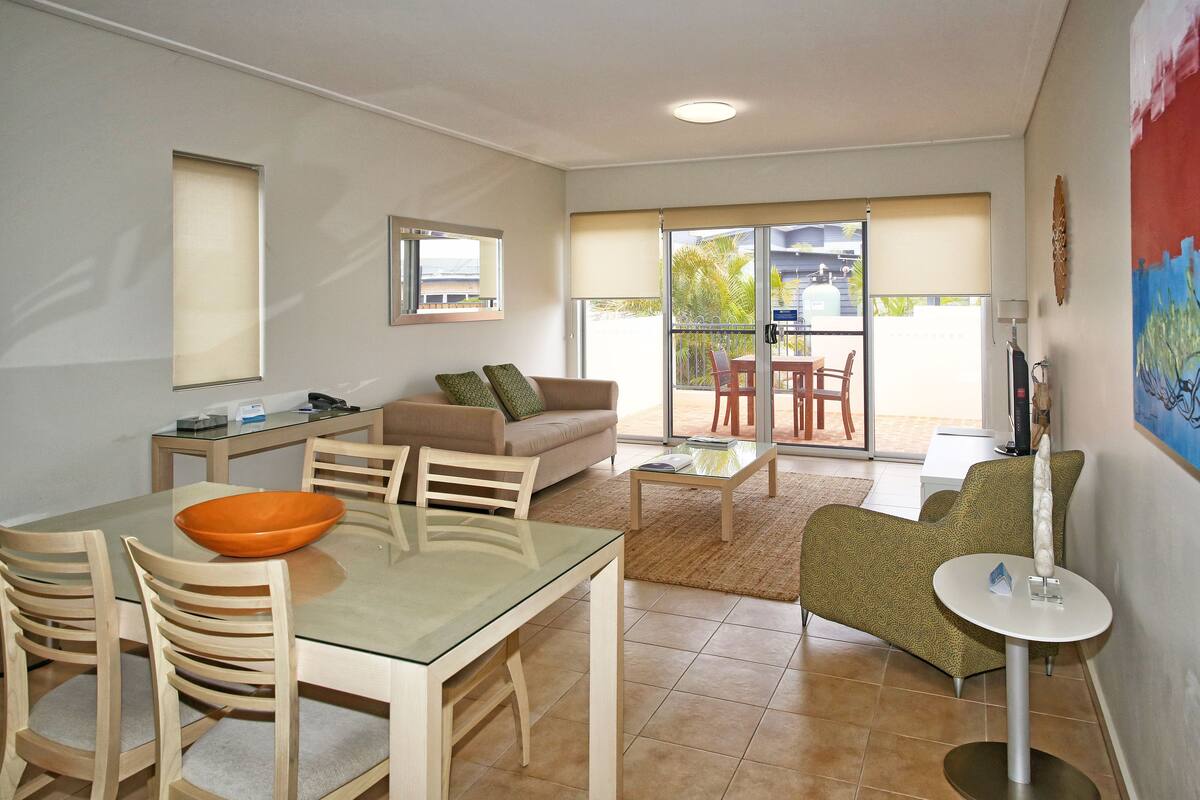 A modern living room with a glass dining table, wooden chairs, a sofa, and a small round table with a lamp.