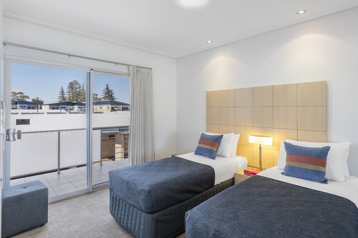 A hotel room with two beds, a balcony, and a view of buildings and trees.