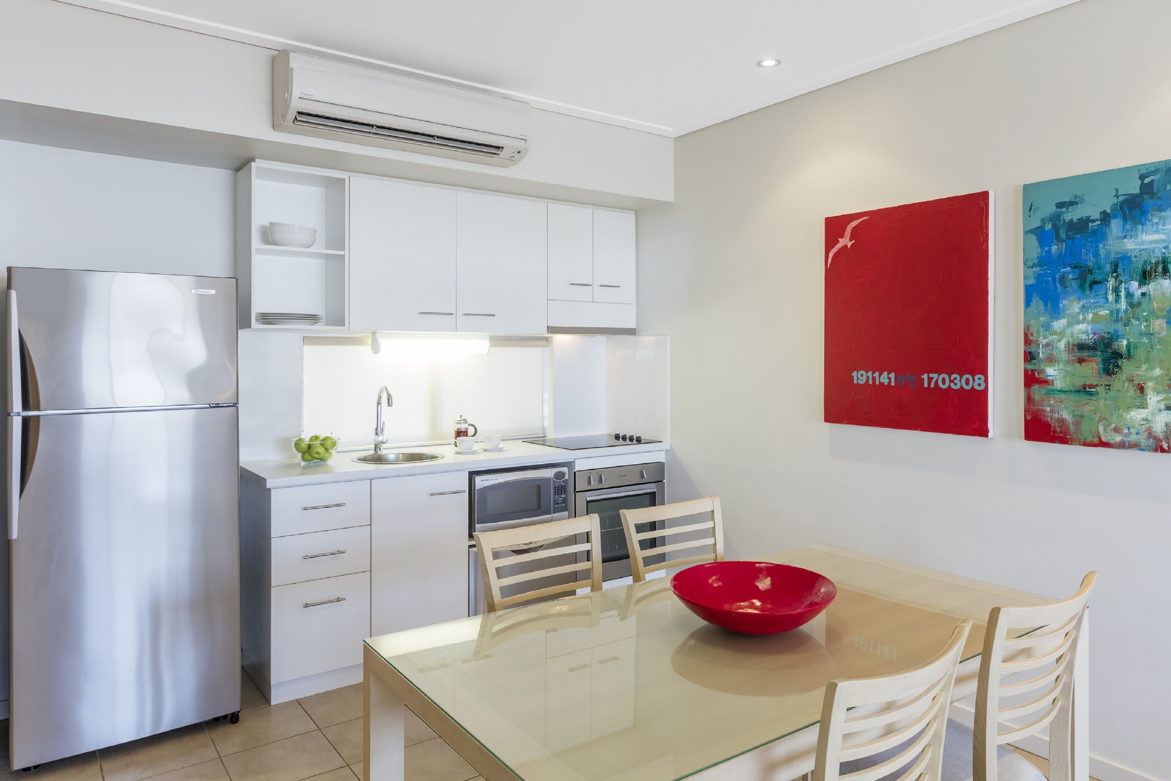 A modern kitchen with white cabinets, stainless steel appliances, and a glass dining table with a red bowl.
