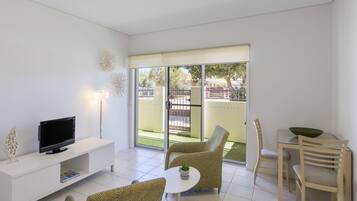 A modern living room with a flat-screen TV on a white console, two patterned armchairs, a small round table with a plant, and a dining area with a green bowl and wooden chairs.