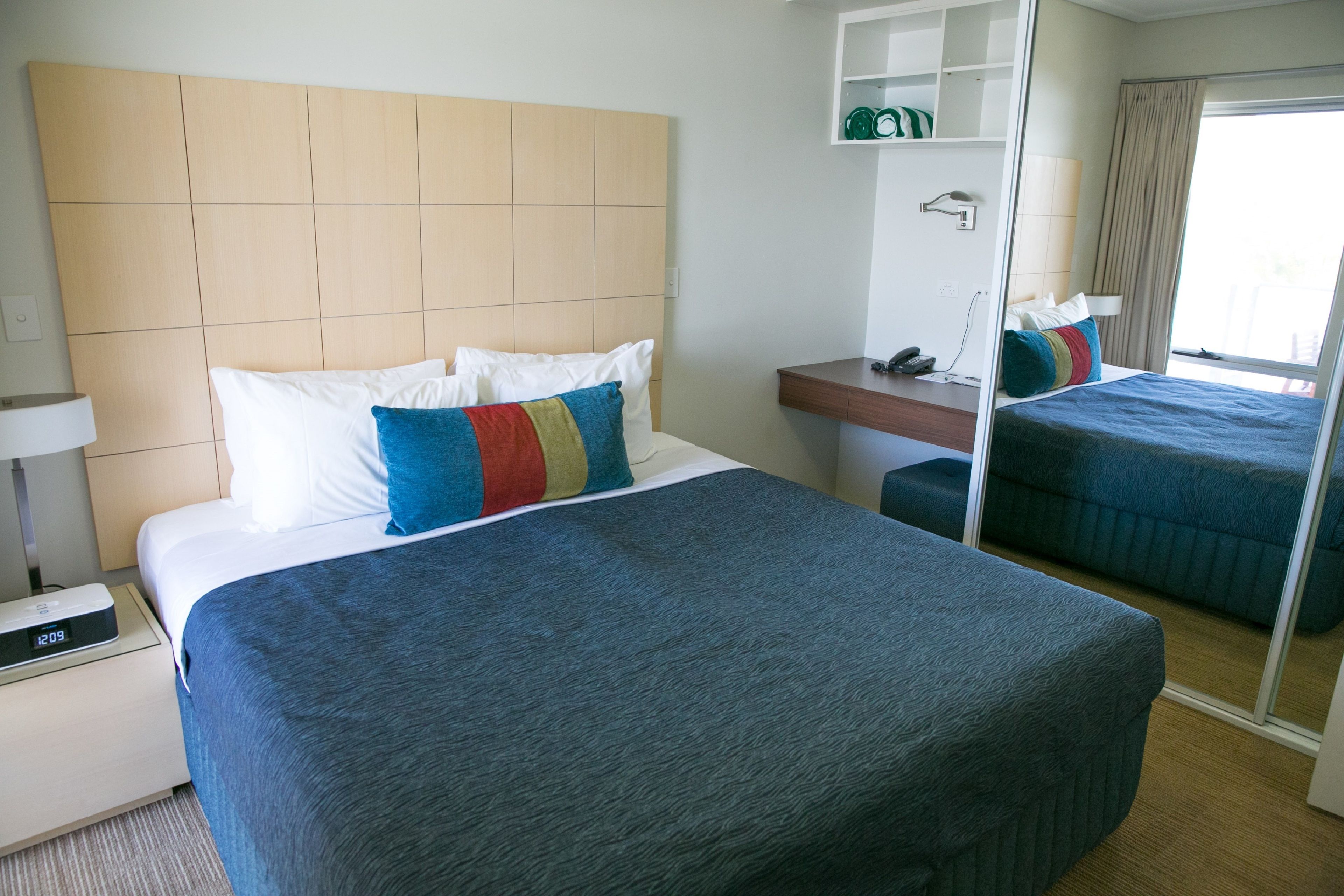 A hotel room with a large bed, a bedside table with a lamp, a small bedside table with a phone, a mirror, and a wardrobe.