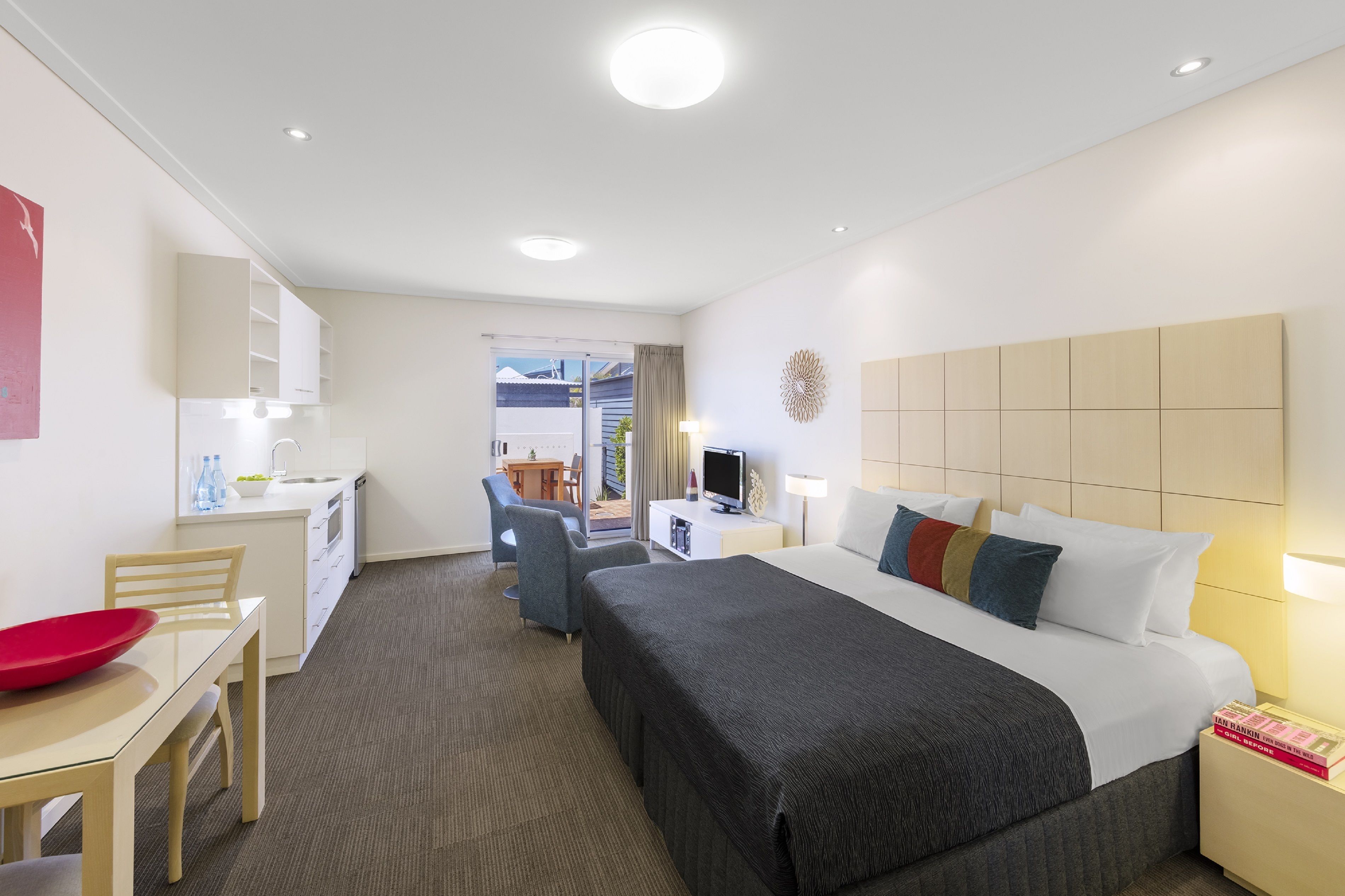 A hotel room with a bed, bedside table, desk, chair, and a small kitchenette.