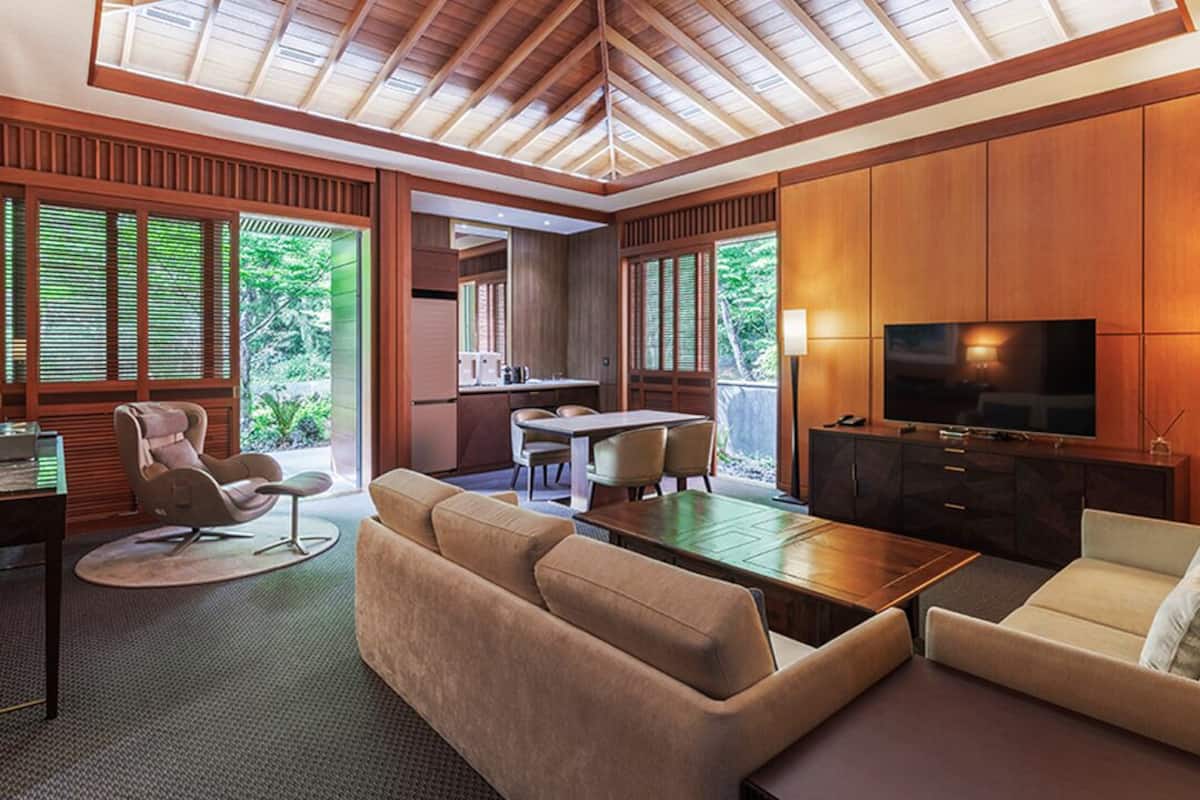A modern living room with a wooden ceiling, a TV, sofas, a dining area, and large windows with blinds.