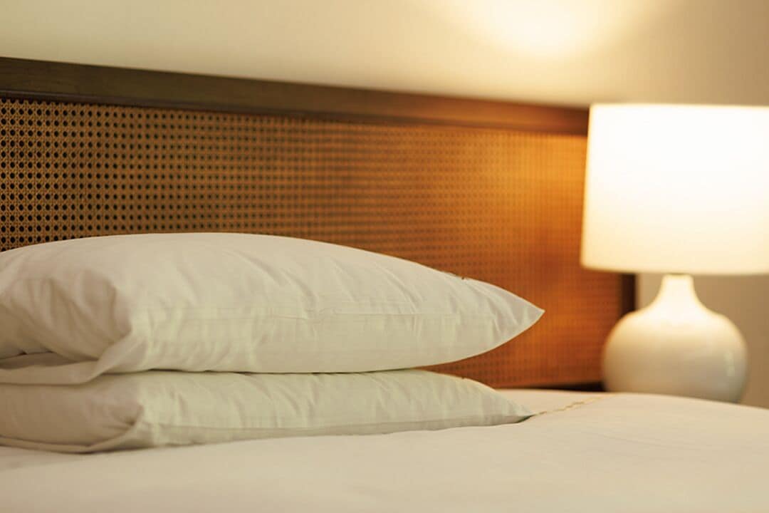 A neatly made bed with white linens and pillows, a wooden headboard, and a white bedside lamp.