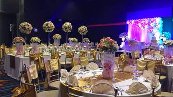 Un salón de banquetes con mesas redondas dispuestas para un evento formal, con sillas doradas, manteles blancos y centros de mesa florales.