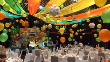 Un salón de banquetes con mesas redondas dispuestas para un evento formal, decorado con telas coloridas y globos.