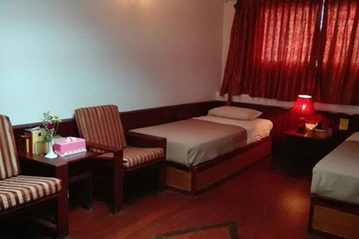 Quarto de hotel com duas camas de solteiro, uma escrivaninha de madeira com cadeira, mesa de cabeceira com abajur e janela com cortinas vermelhas.