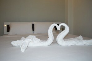 A bed with white linens and pillows, and two towels arranged in the shape of swans forming a heart. - Nan Treasure (Phu Phiang)