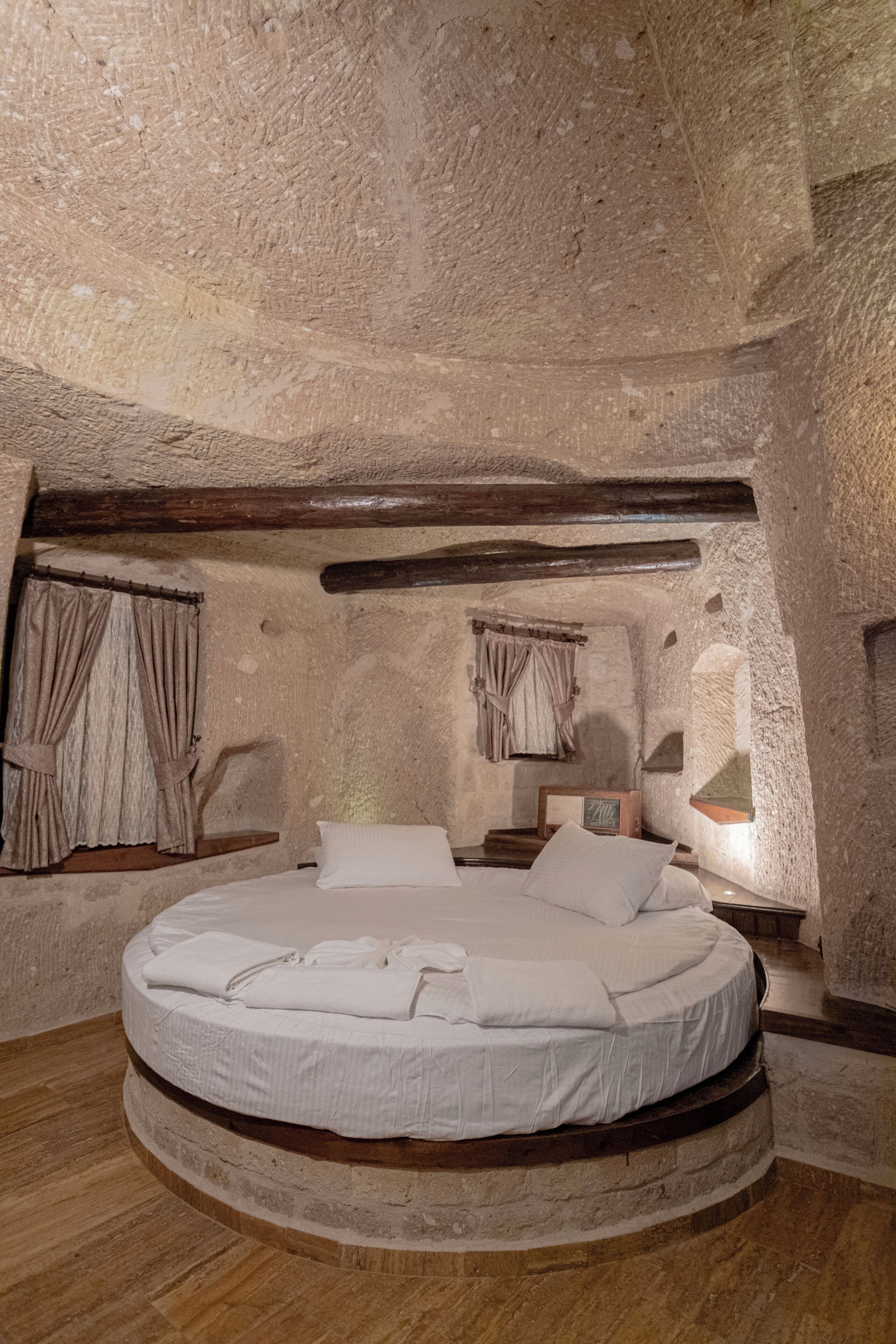 Una cama redonda en una habitación rústica con paredes de piedra, vigas de madera y un pequeño ventanal con cortinas.