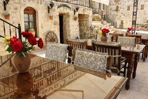 Un área de comedor al aire libre con paredes de piedra, mesas y sillas de madera. Hay flores en macetas sobre las mesas.
