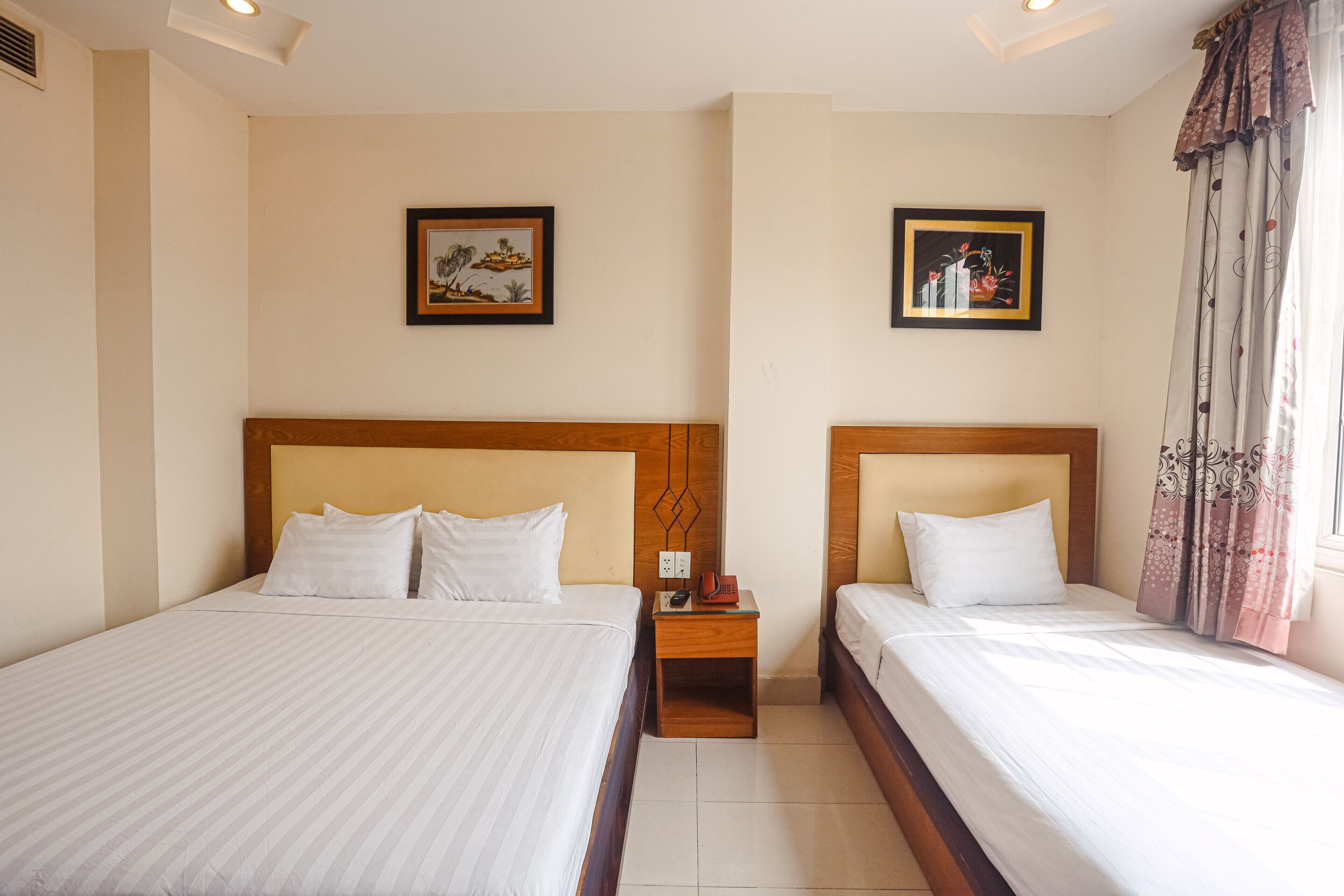 Een hotelkamer met twee bedden, elk met een houten hoofdbord en witte beddengoed. Er hangen twee ingelijste foto's aan de muur, een nachtkastje met een telefoon en een rood object tussen de bedden, en een raam met gordijnen.