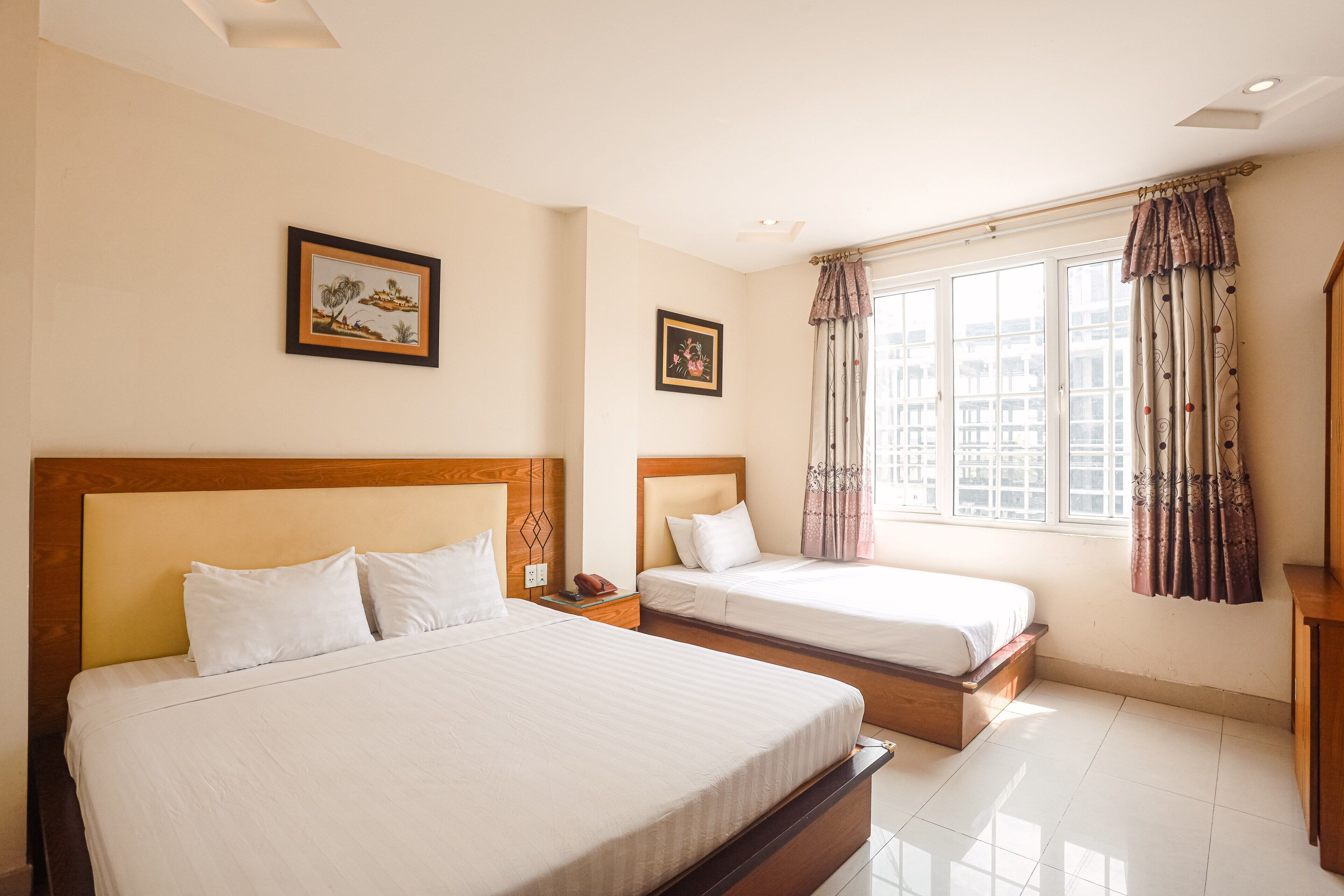 A hotel room with two beds, a large window with curtains, and framed artwork on the walls.